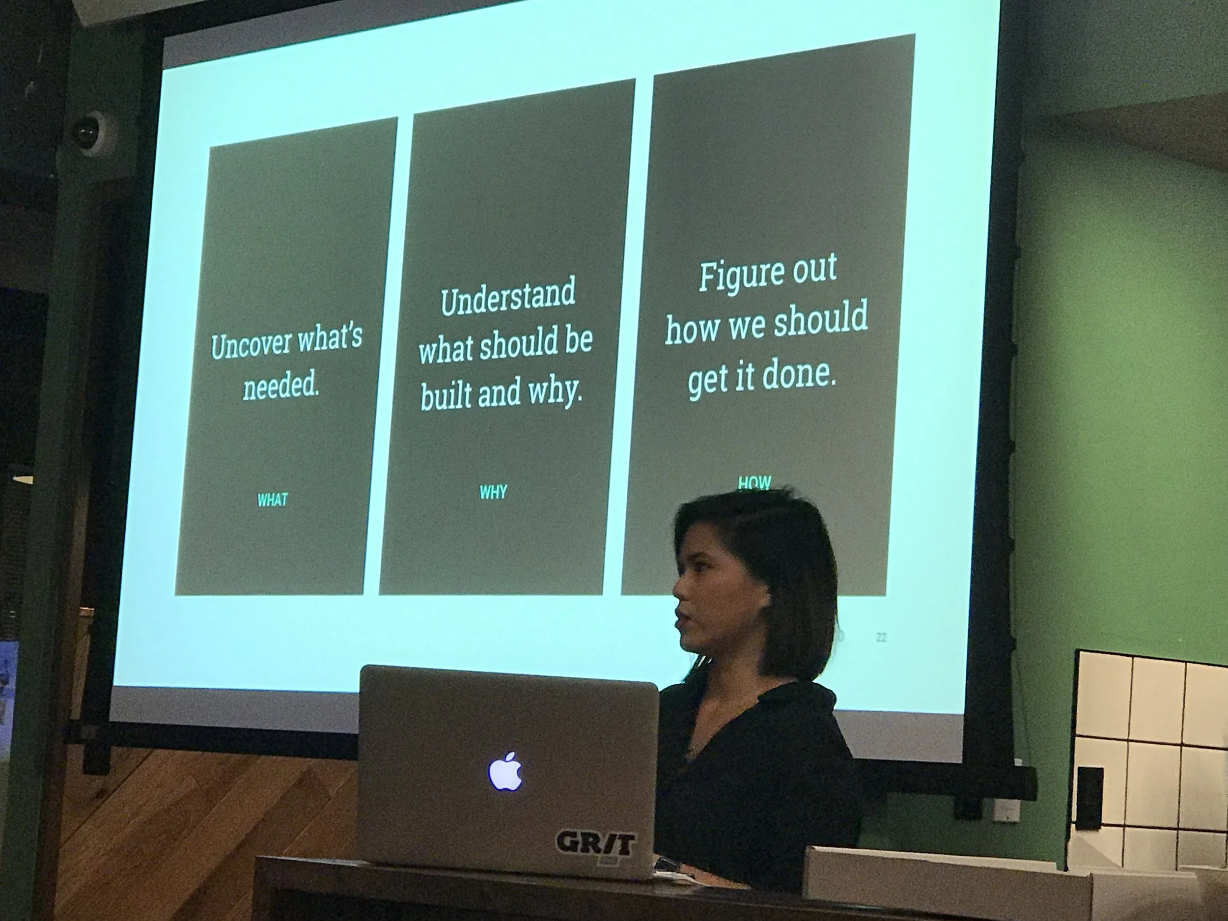 A presentation slide with three sections titled 'What', 'Why', and 'How'. The content reads: 'Uncover what's needed.', 'Understand what should be built and why.', and 'Figure out how we should get it done.' In the foreground, a woman is sitting in fr
