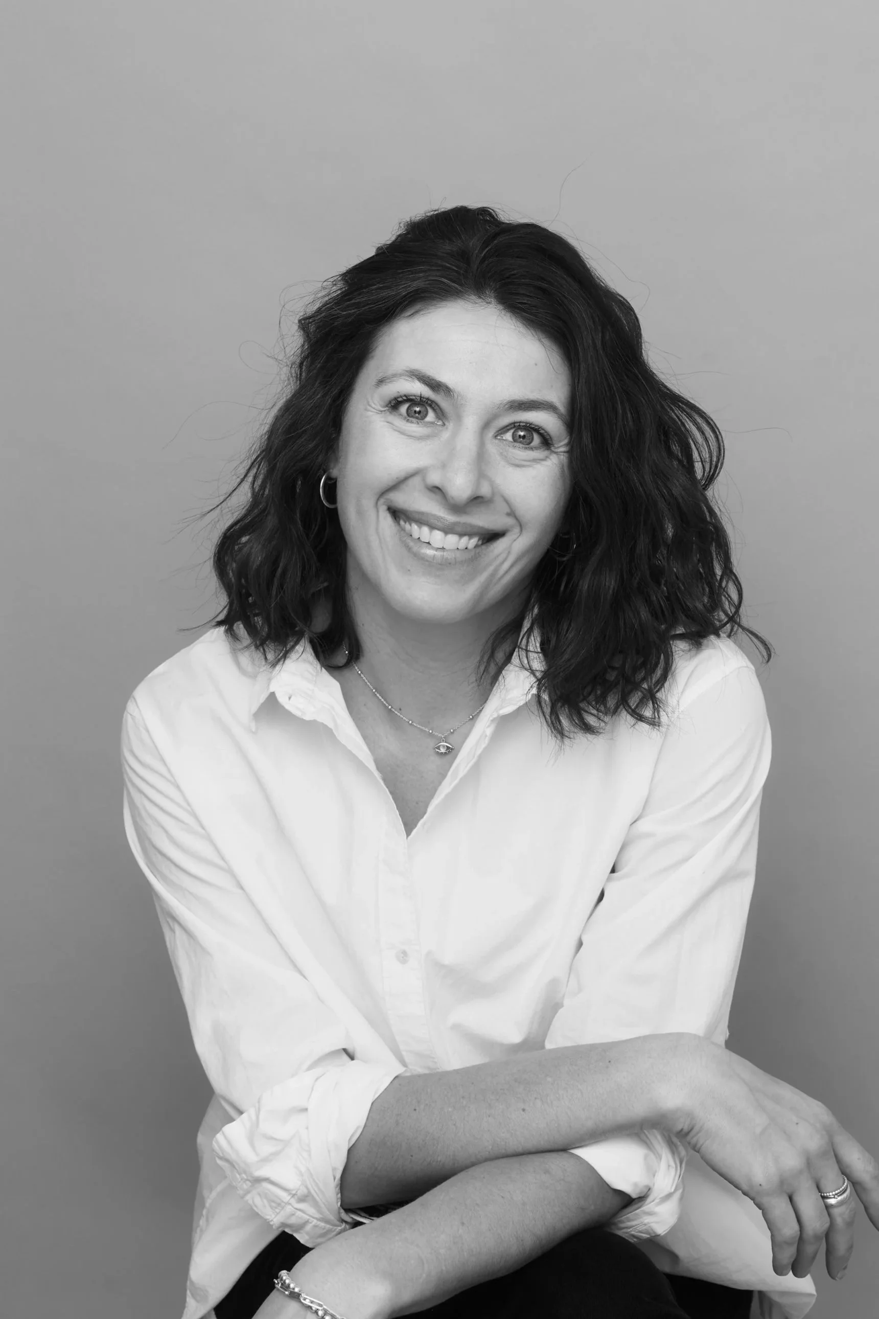 An image of Caley Bell-Pasht smiling, wearing a white shirt, sitting with her arms crossed and resting on her knees, against a plain background.