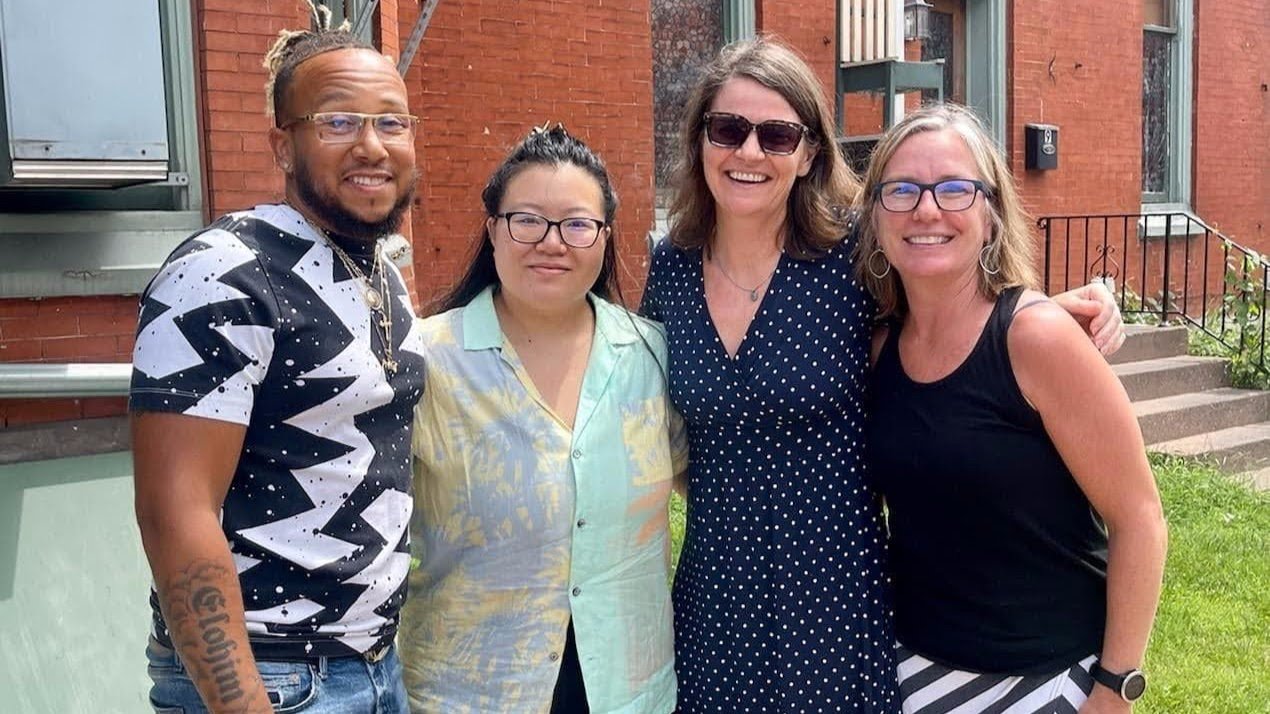 Four women and one man smiling outdoors in front of a brick building, posing together for a photo.