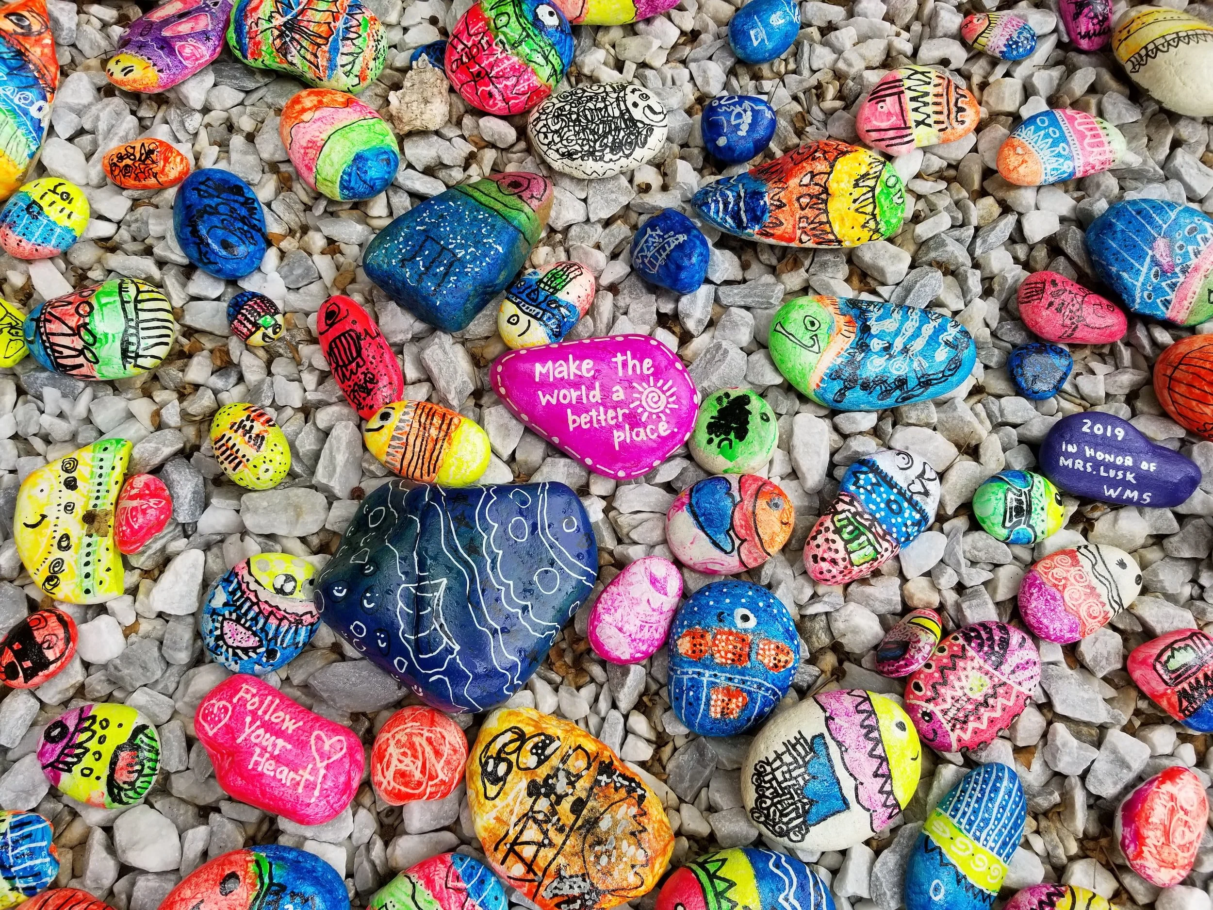Colorful painted rocks of various sizes scattered on a gray gravel surface, with some rocks featuring drawings, patterns, and messages such as 'Make the world a better place' and 'Follow your heart' in bright colors.