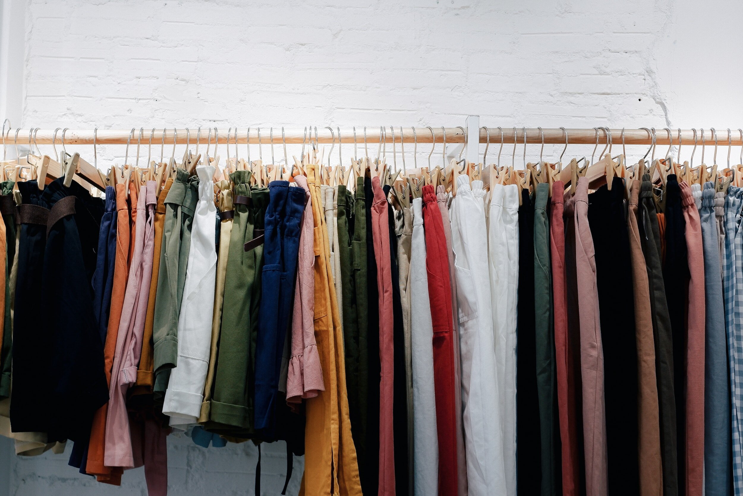 A clothing rack with various colorful pants hanging on wooden hangers against a white brick wall.