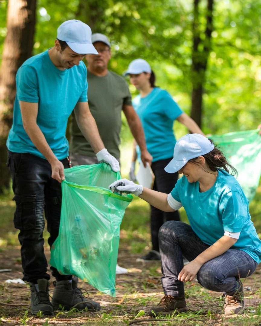 Group of volunteers in a forest collecting trash, wearing gloves and caps, with one person holding a green trash bag.