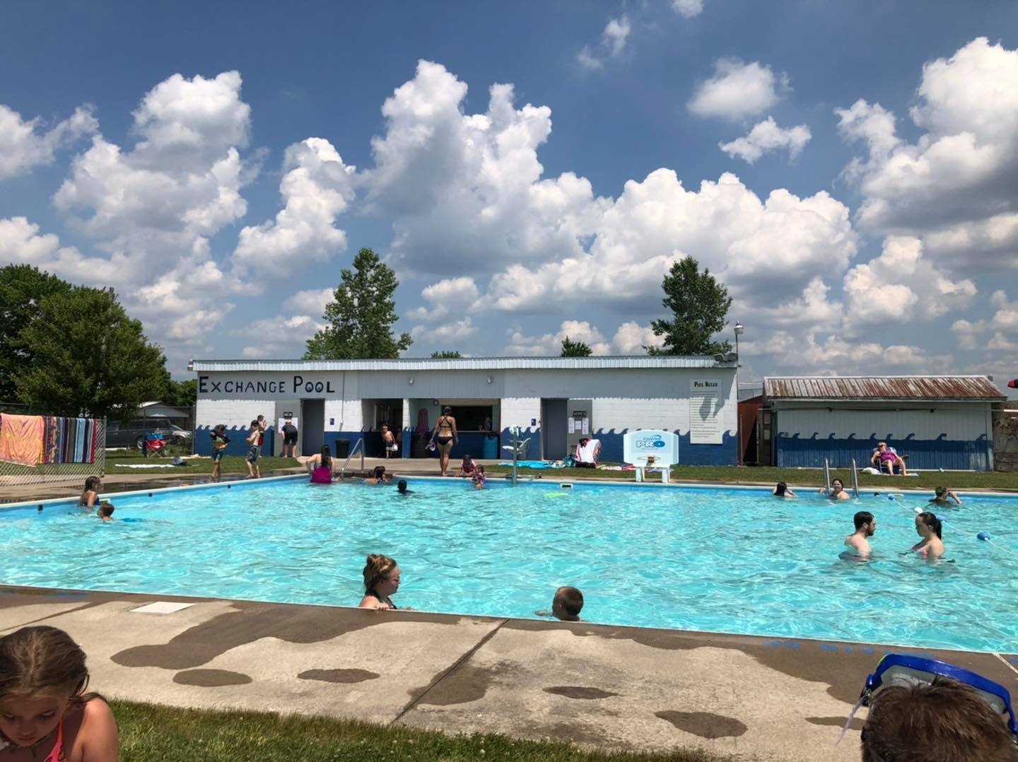 People enjoying swimming in an outdoor pool with a sign that reads 'Exchange Pool.' There are children and adults in the water, with some lounging on the poolside. The background features a white building with the sign and blue skies with scattered clouds.