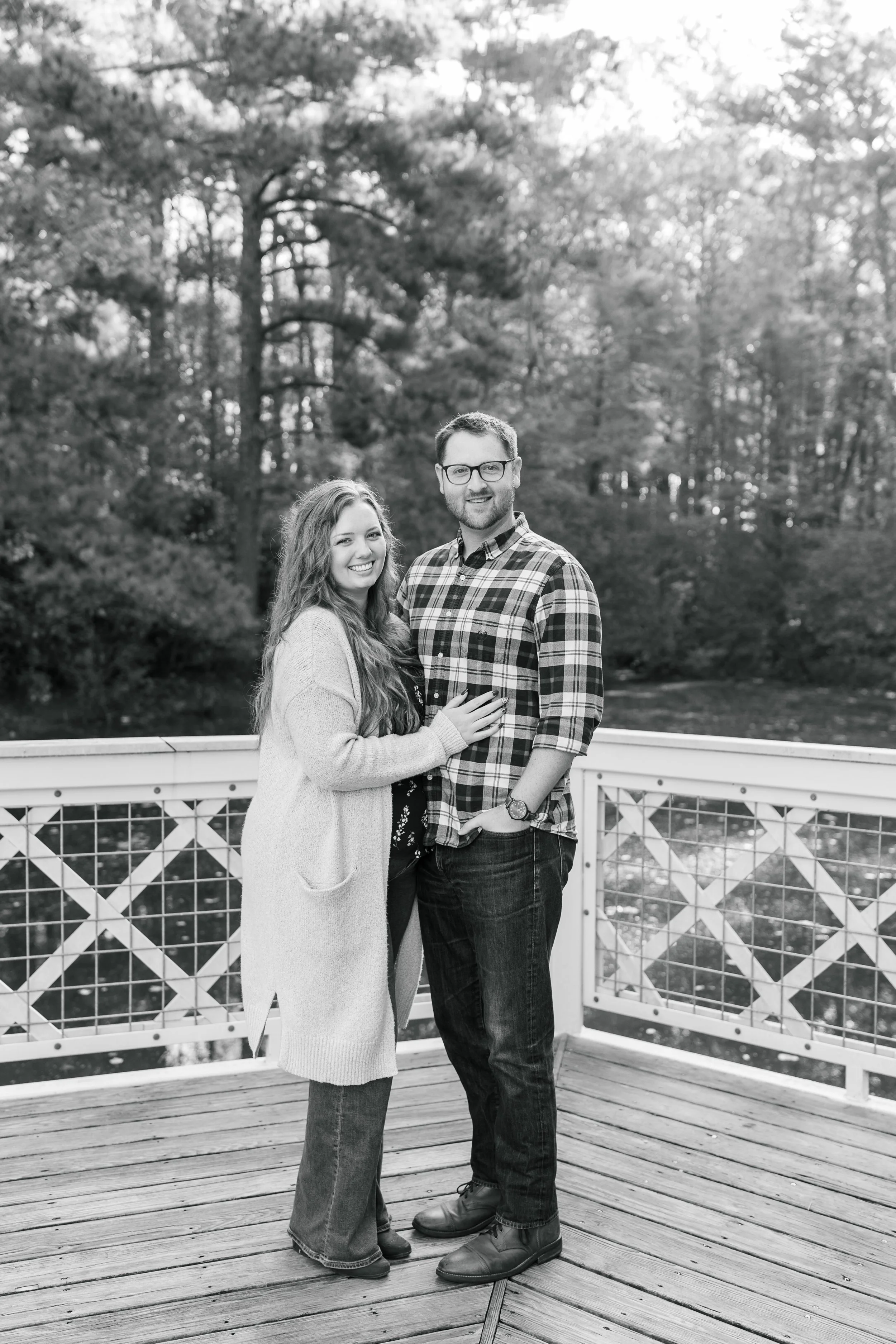 A smiling young woman with long hair and a man with glasses and a beard, standing on a wooden deck with a white railing, surrounded by trees in a natural outdoor setting.