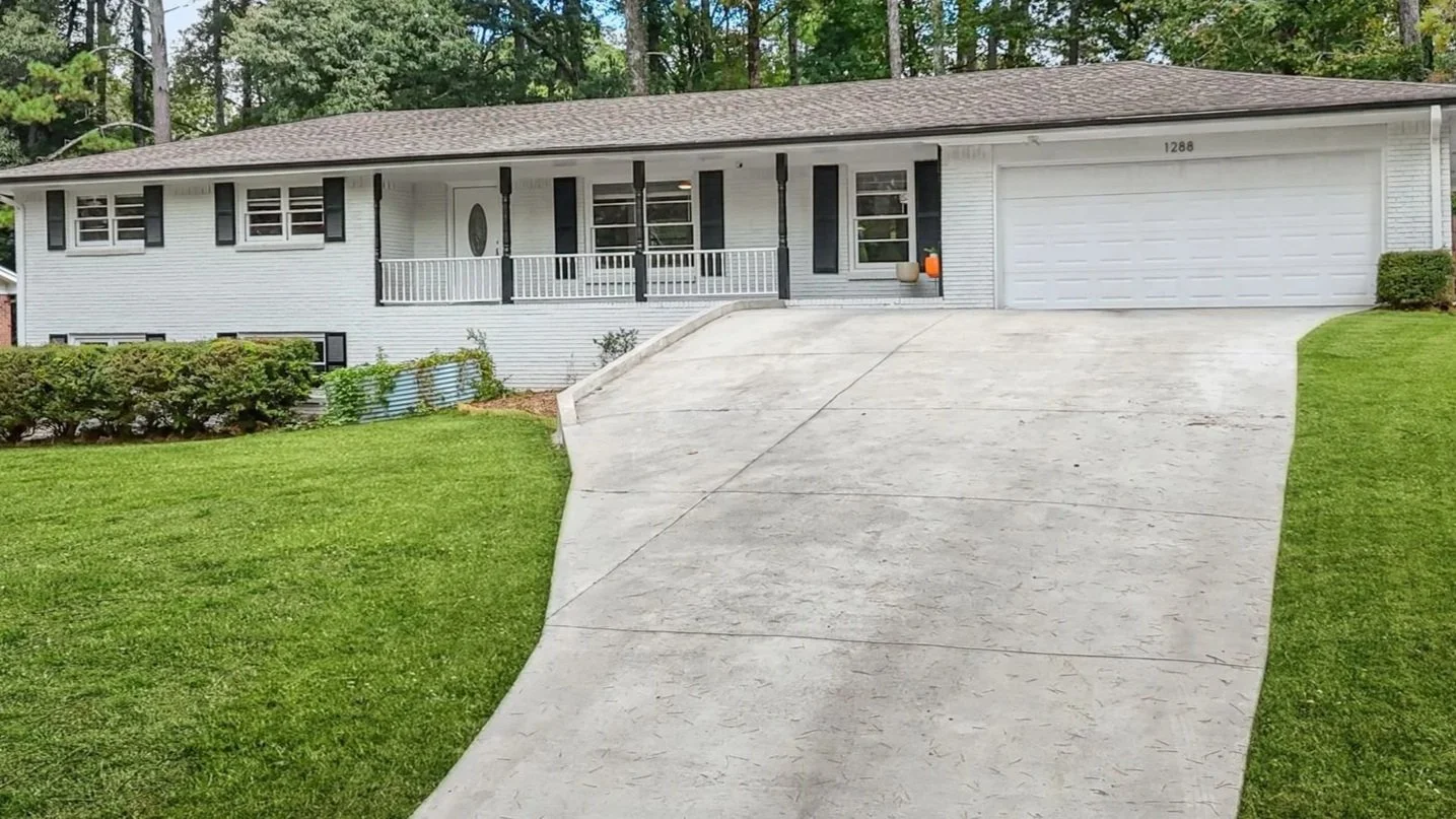 A white ranch-on-a-basement house with a two-car garage, a concrete driveway, and a front porch, surrounded by a green lawn and trees in the background.
