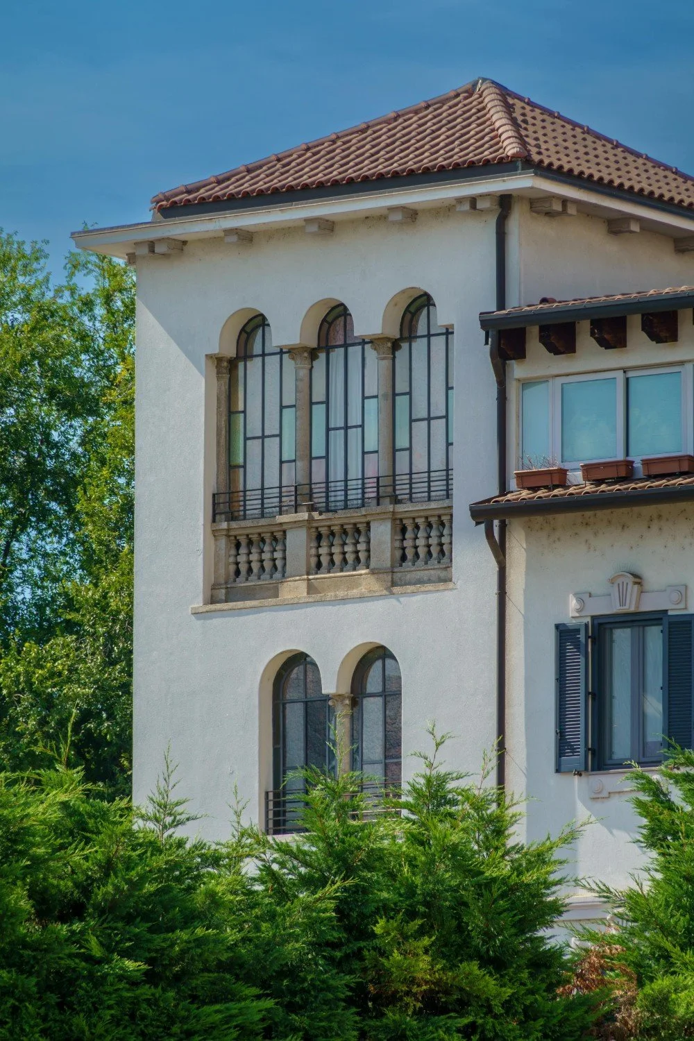 A multi-story house with a red-tiled roof, arched windows with decorative columns, and a small balcony. The house has white stucco walls, black shutters, and is surrounded by green shrubbery and trees.