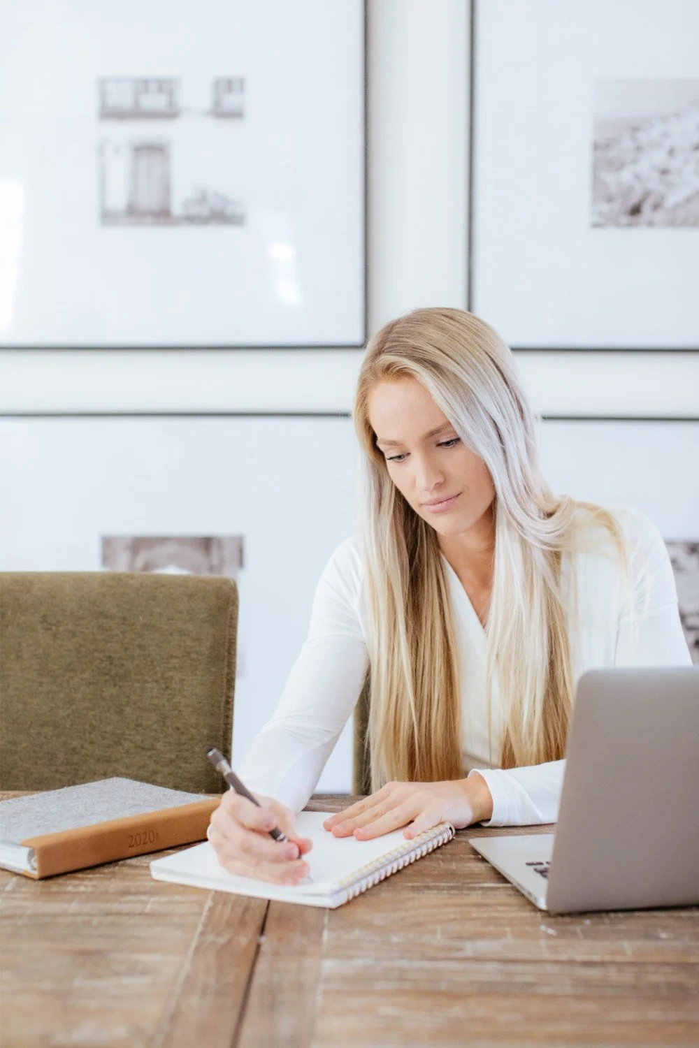 A woman with long blonde hair writes in a spiral notebook at a wooden table, with a laptop and a planner nearby.