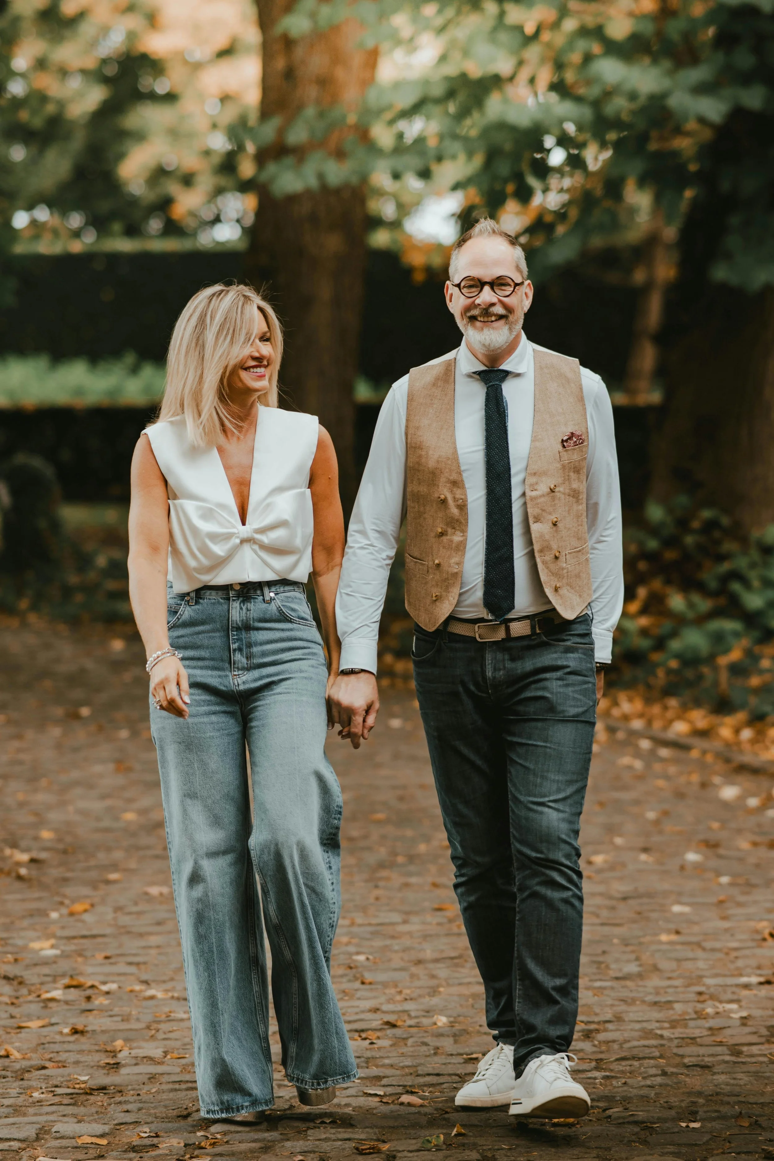 A man and woman walking hand-in-hand outdoors on a tree-lined path, smiling and enjoying each other's company.