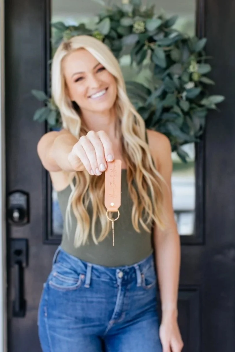 A woman with long blonde hair, smiling and holding up a keychain with a key, standing in front of a door with greenery in the background.