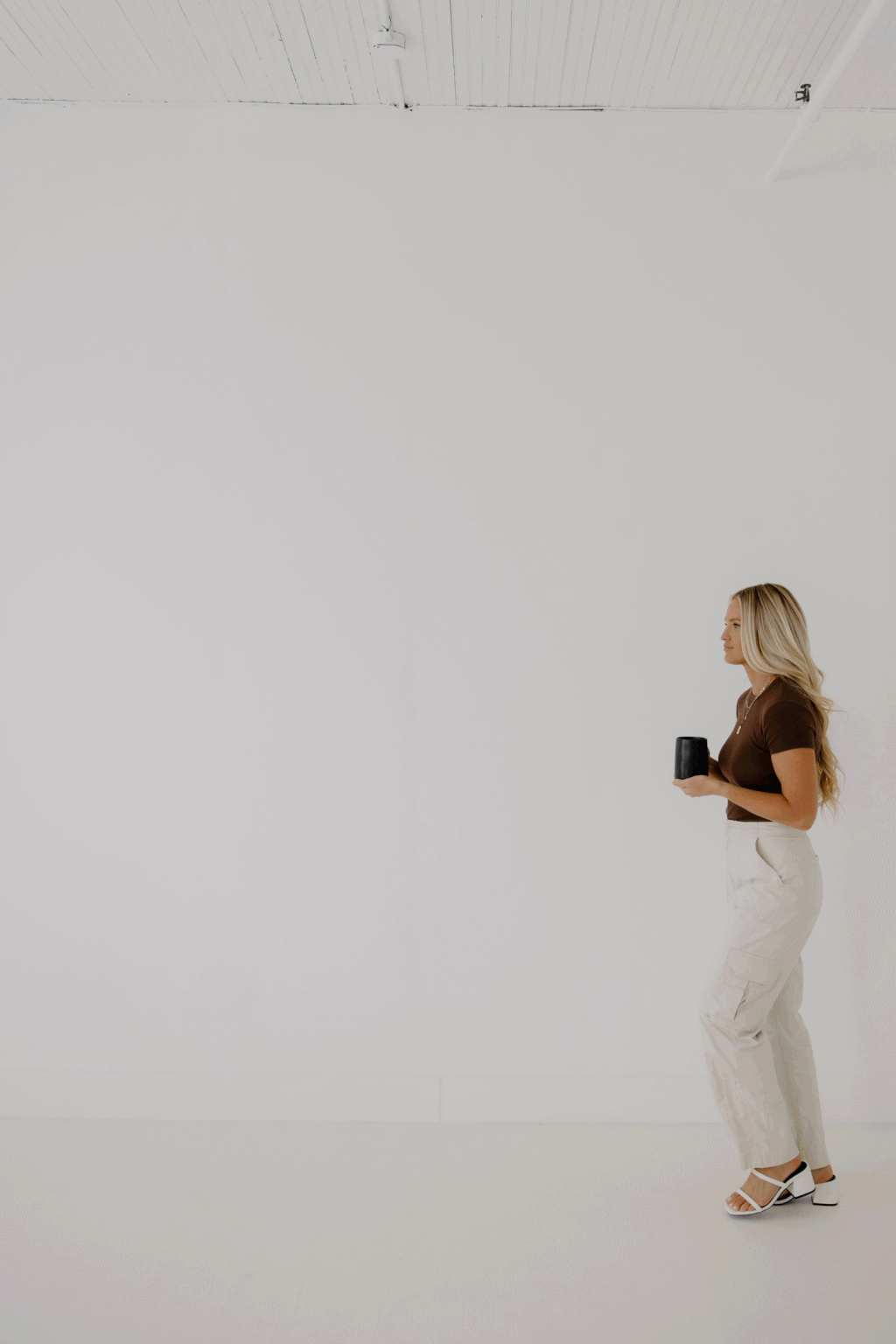 Woman with long blonde hair holding a black mug, standing sideways in a minimalist white room.