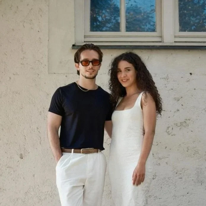 A man with dark hair, sunglasses, and light skin wearing a black shirt and white pants standing next to a woman with curly dark hair and light skin wearing a white dress, outdoors in front of a white wall with a window.
