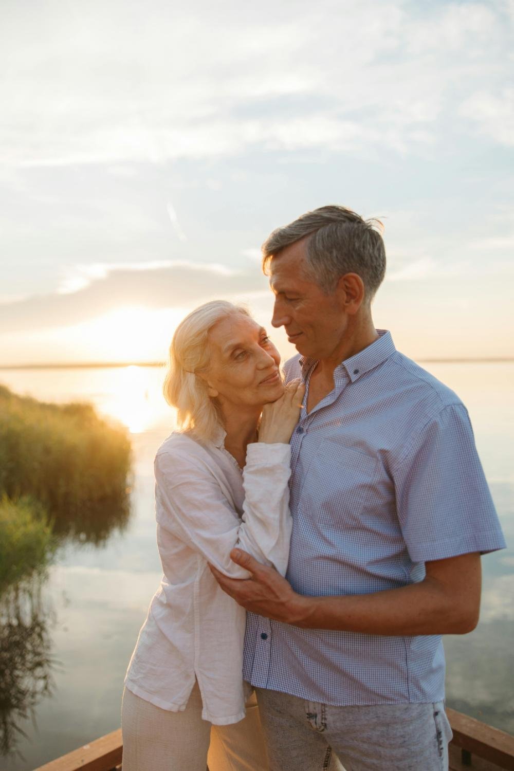 An elderly couple sharing a loving moment by a body of water during sunset, embracing each other gently.