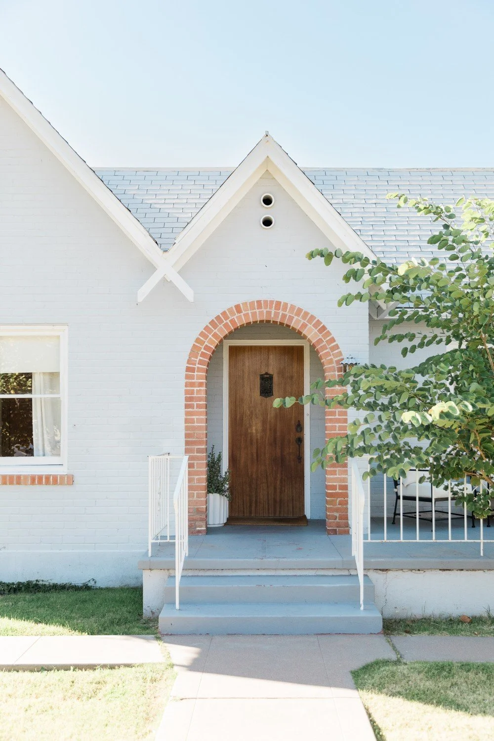 White house with brick arch around wooden front door, stairs with white railings, green tree on right, window on left, and a blue sky