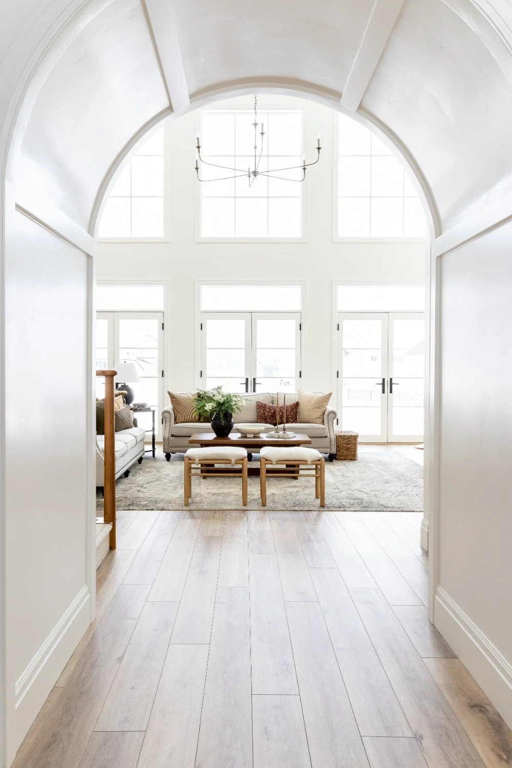 Bright living room with large windows, high ceiling, and minimalist decor. Includes a beige sofa, wooden coffee table, and matching stools.