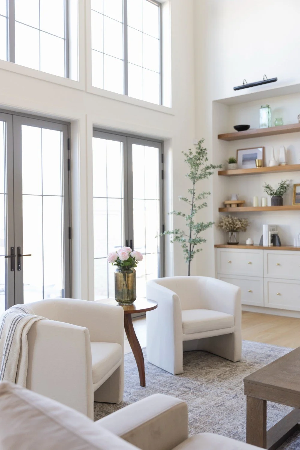 Bright living room with large windows, a double door, white armchairs, a wooden side table with pink flowers, and open wall shelves with decorative items.