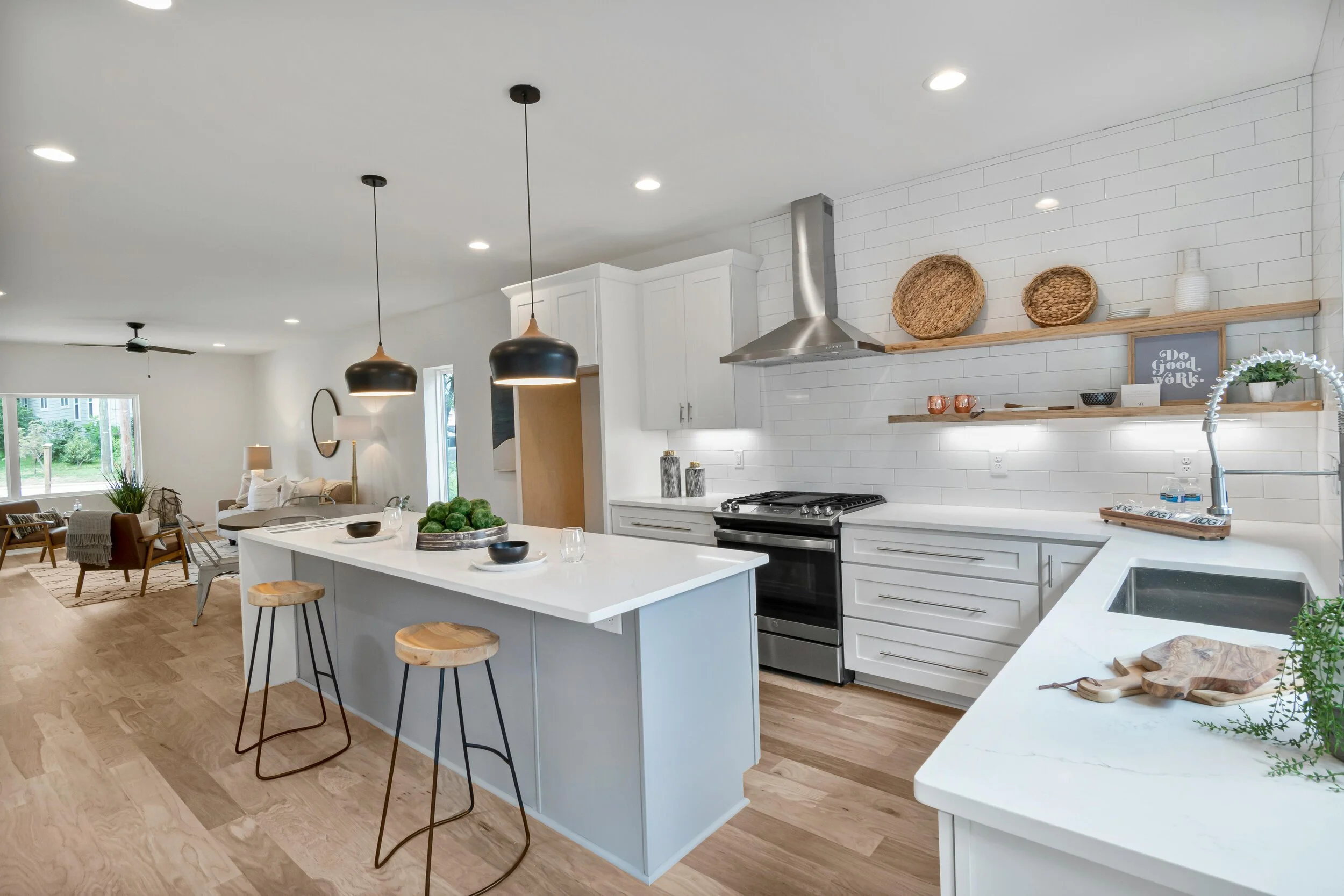 Modern open-concept kitchen with white cabinets, a kitchen island, black pendant lights, wooden shelves, and natural decor, blending into a living area with large windows and seating.