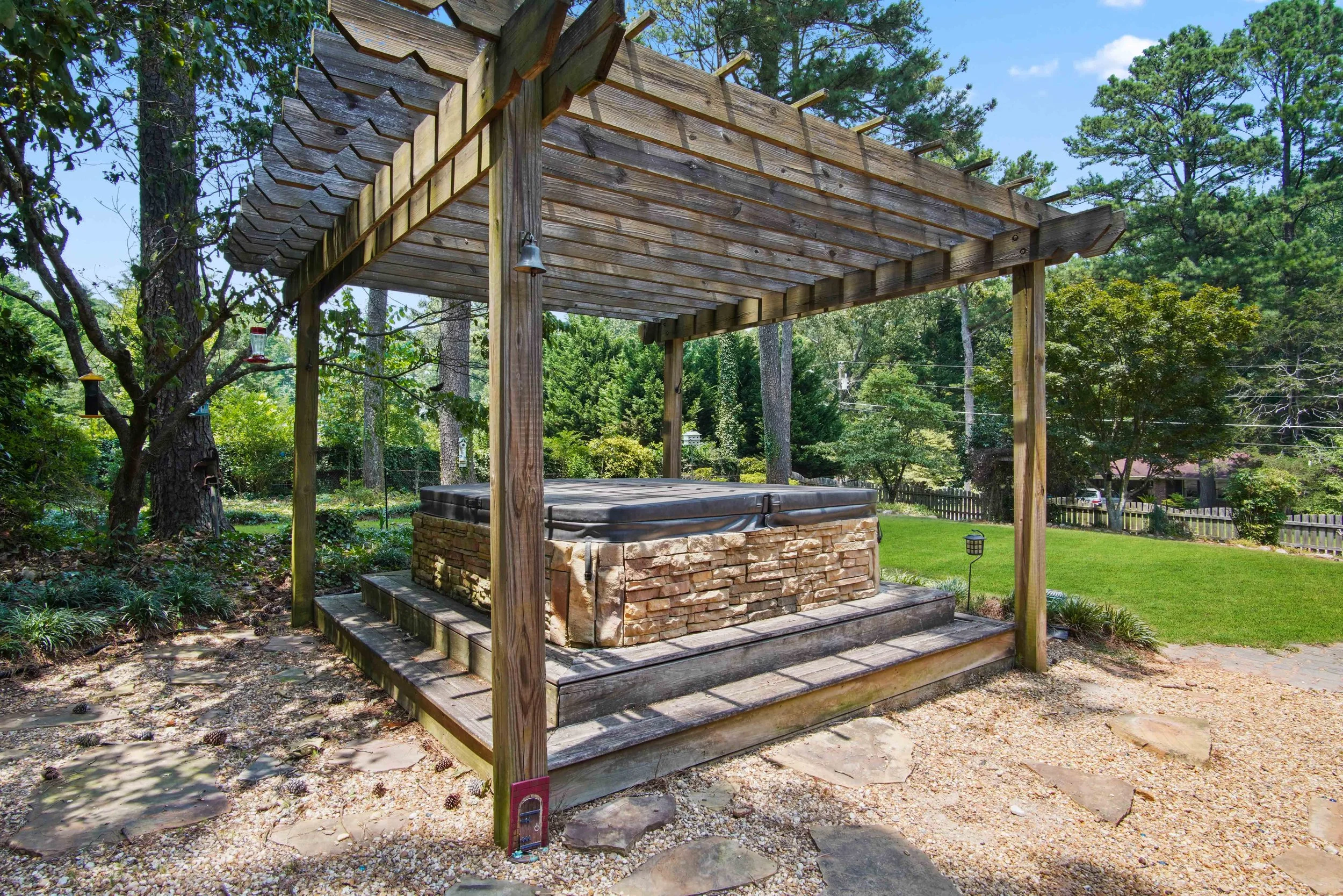 Hot tub with a stone facade beneath a wooden pergola in a backyard, surrounded by trees and a green lawn.