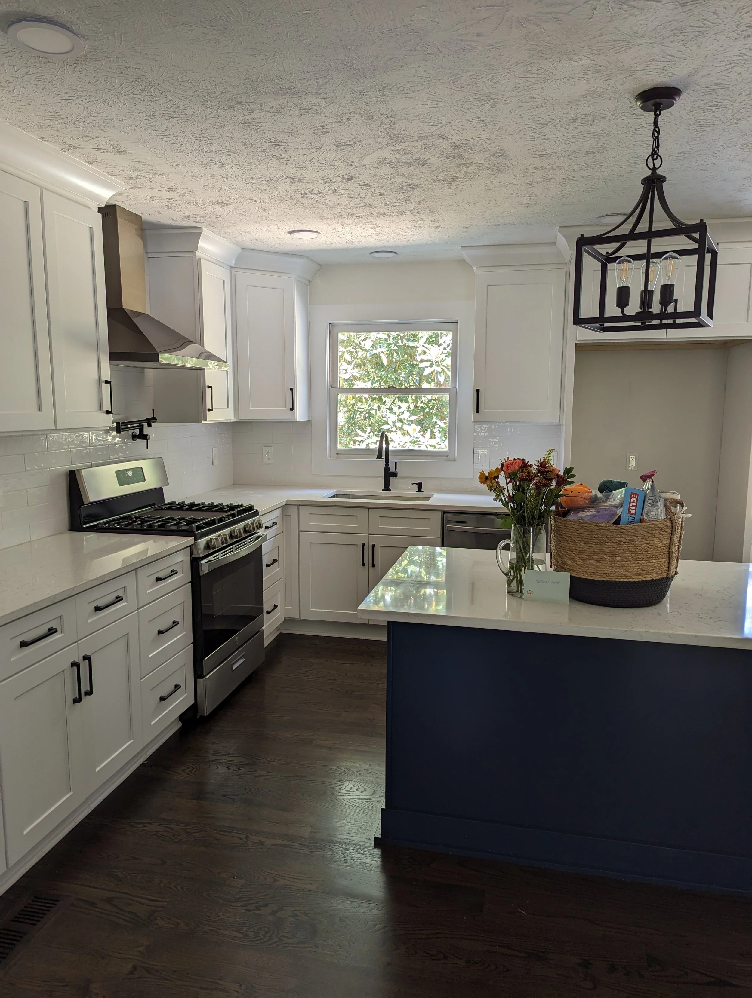 Modern kitchen with white cabinets, black hardware, stainless steel stove, window at the sink, dark hardwood floors, countertop with flowers and a basket of groceries.