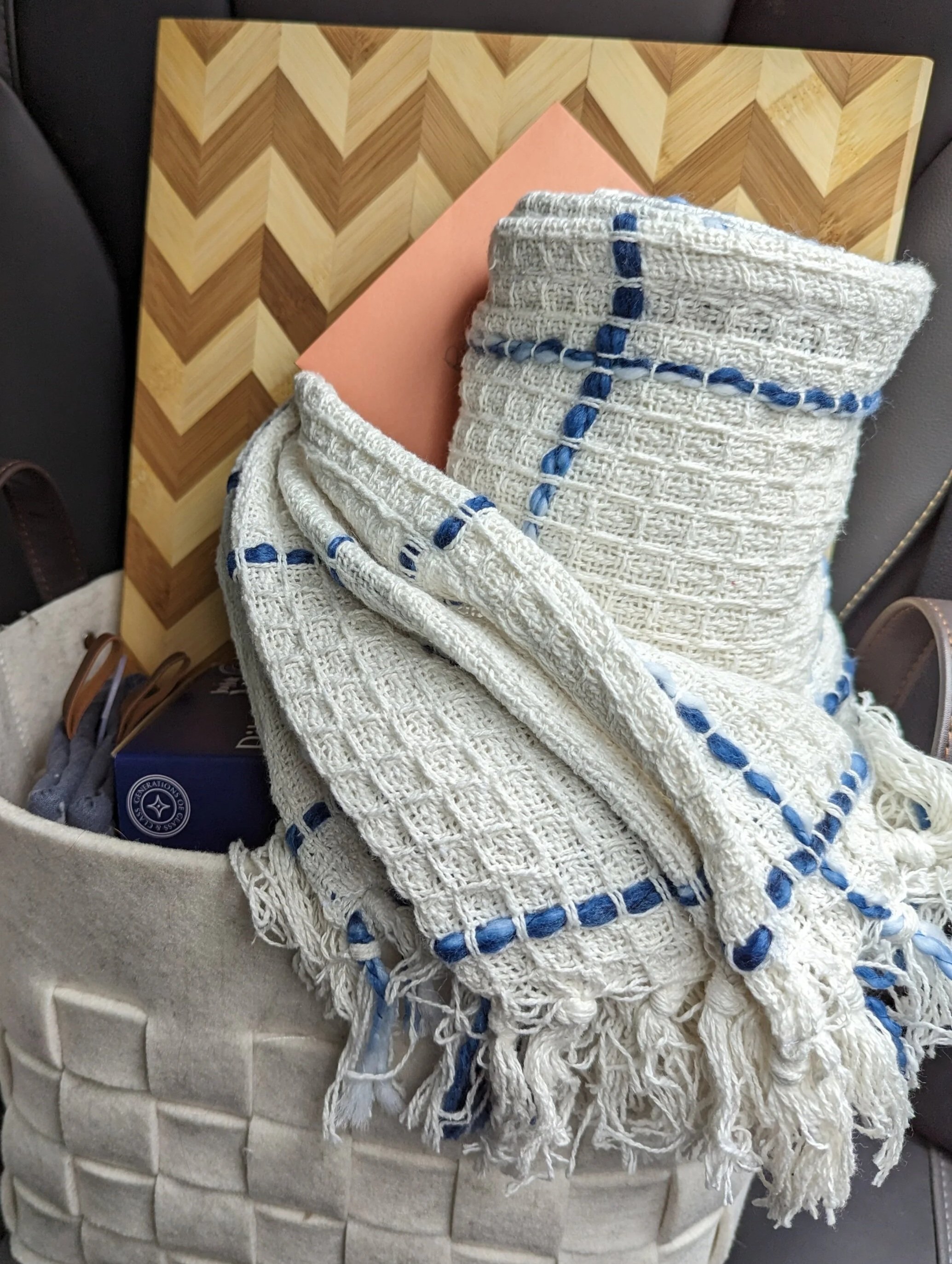 A beige woven basket filled with a white textured throw blanket with blue accents, a pink envelope, and a box of aromatic tea, all placed on a dark chair.