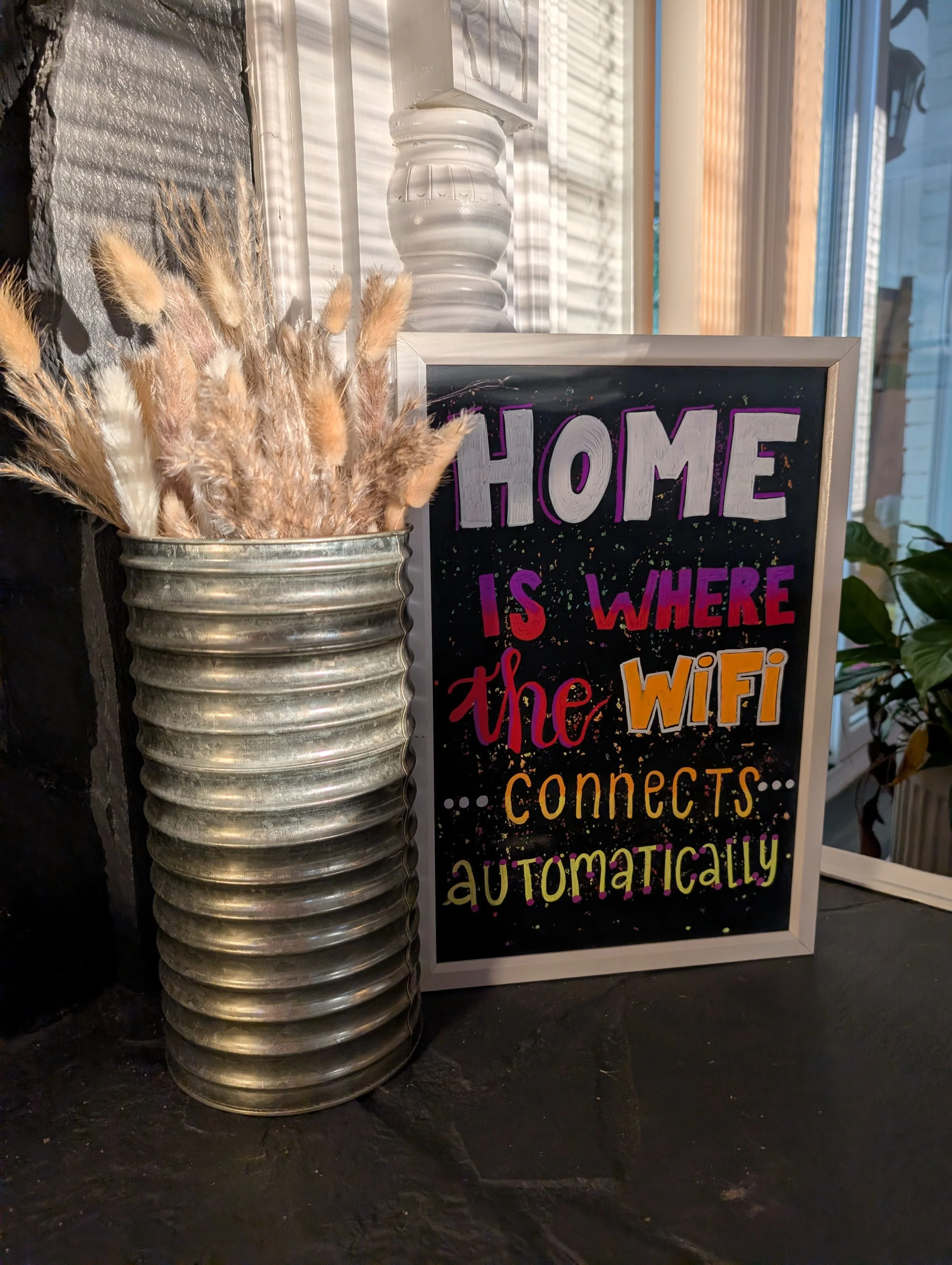 Decorative sign with the text 'Home is where the WiFi connects automatically' placed on a dark surface next to a silver vase filled with dried decorative grasses, near a window with sunlight casting shadows.