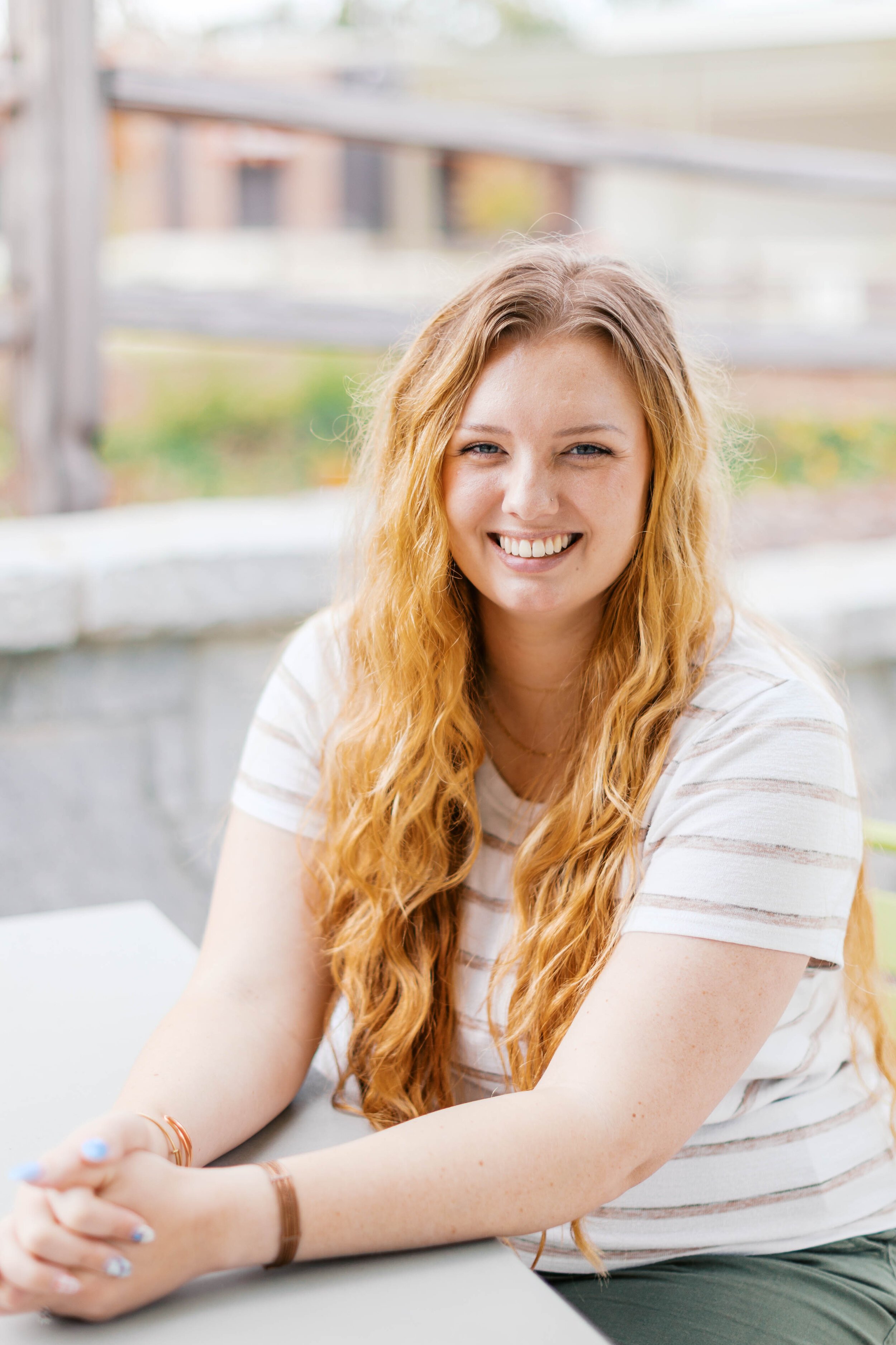 A young woman with long, wavy red hair smiling, sitting at a table outside.