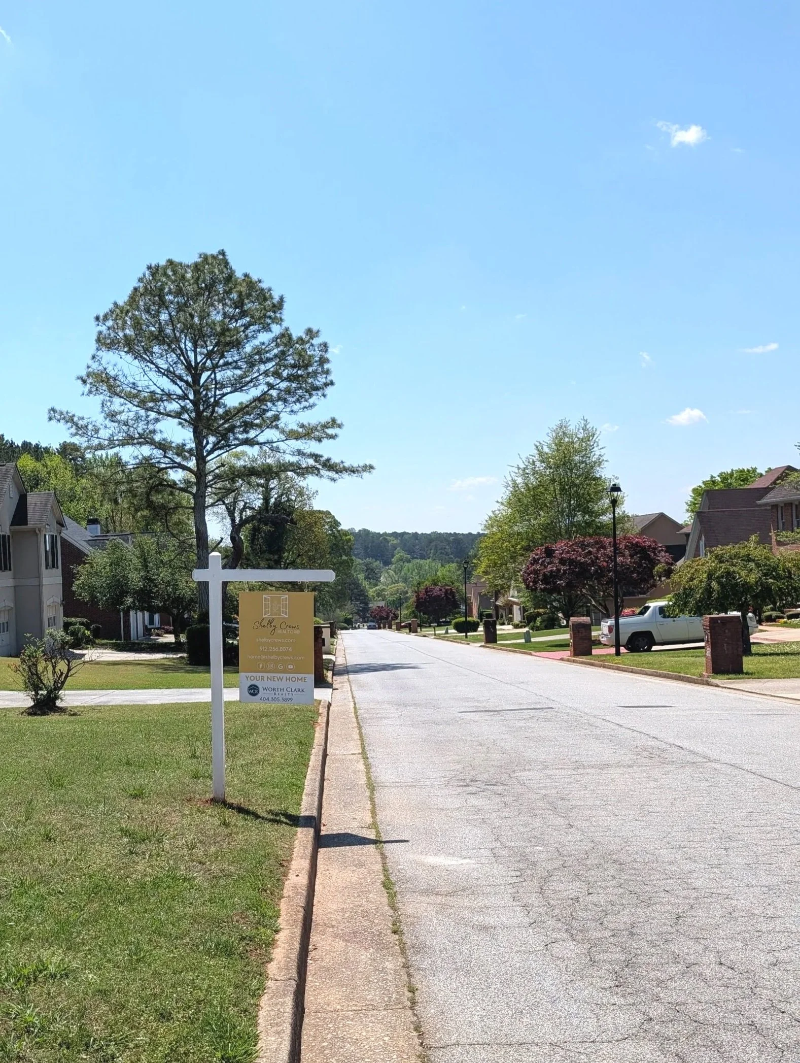 A quiet suburban street with houses, trees, parked cars, and a sign in the front yard advertising a home for sale, under a clear blue sky.