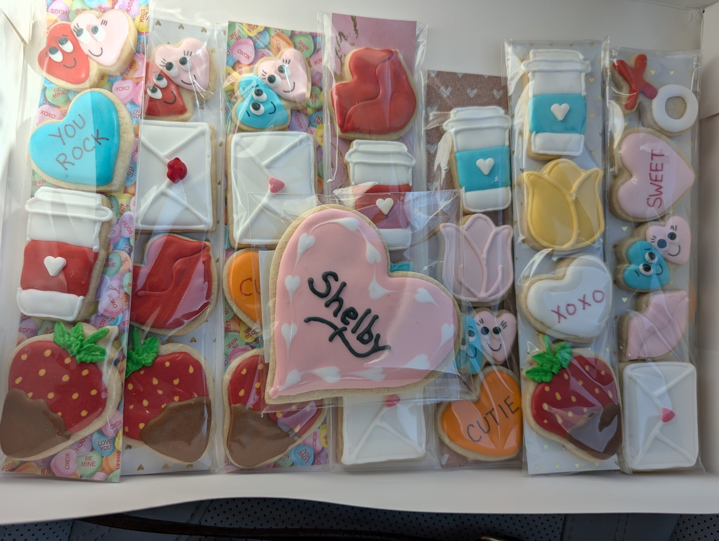 Assorted decorated cookies wrapped in plastic, featuring hearts, strawberries, flowers, and messages like "You Rock," "Shelby," "Cute," "XOXO," and "Sweet."