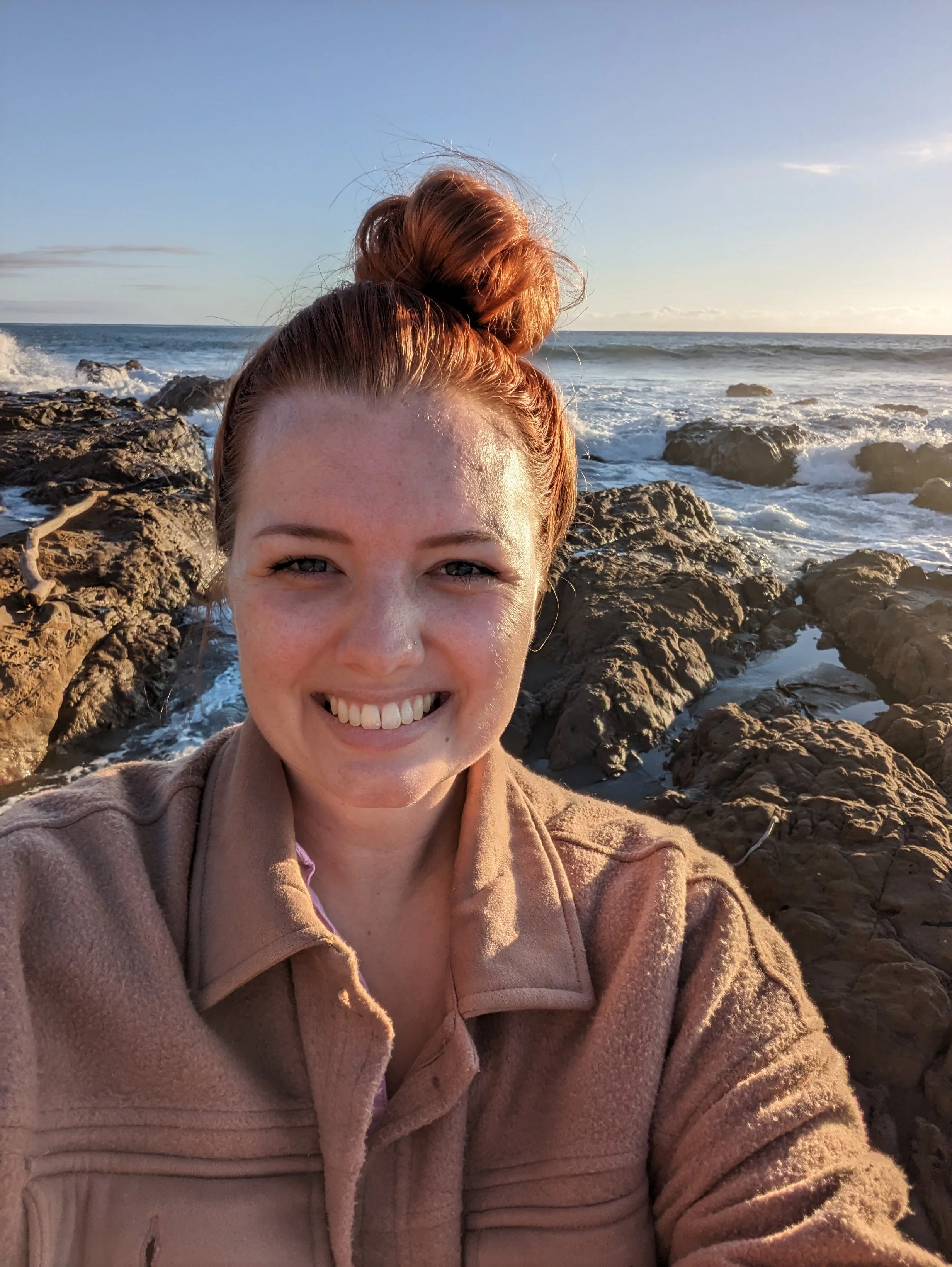 A woman with red hair styled in a bun, smiling at the camera, with rocks and ocean waves in the background during sunset.
