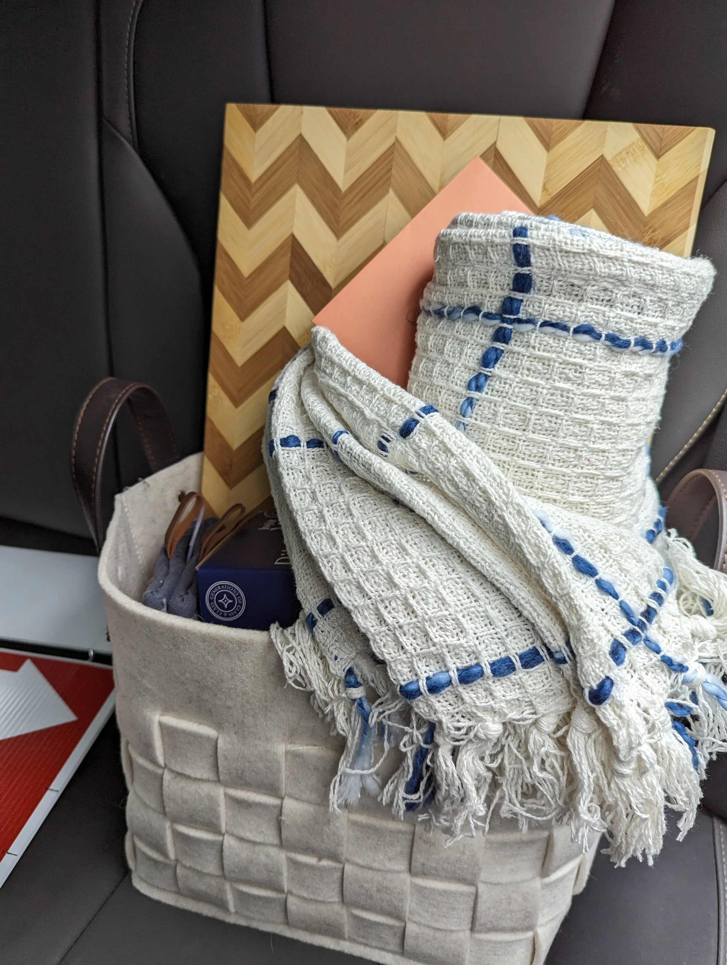 A beige woven basket filled with clothes, a hardcover book with an orange cover, a blanket with blue stitched circles, a navy box of chocolates, and a pair of scissors, placed on the back seat of a car.