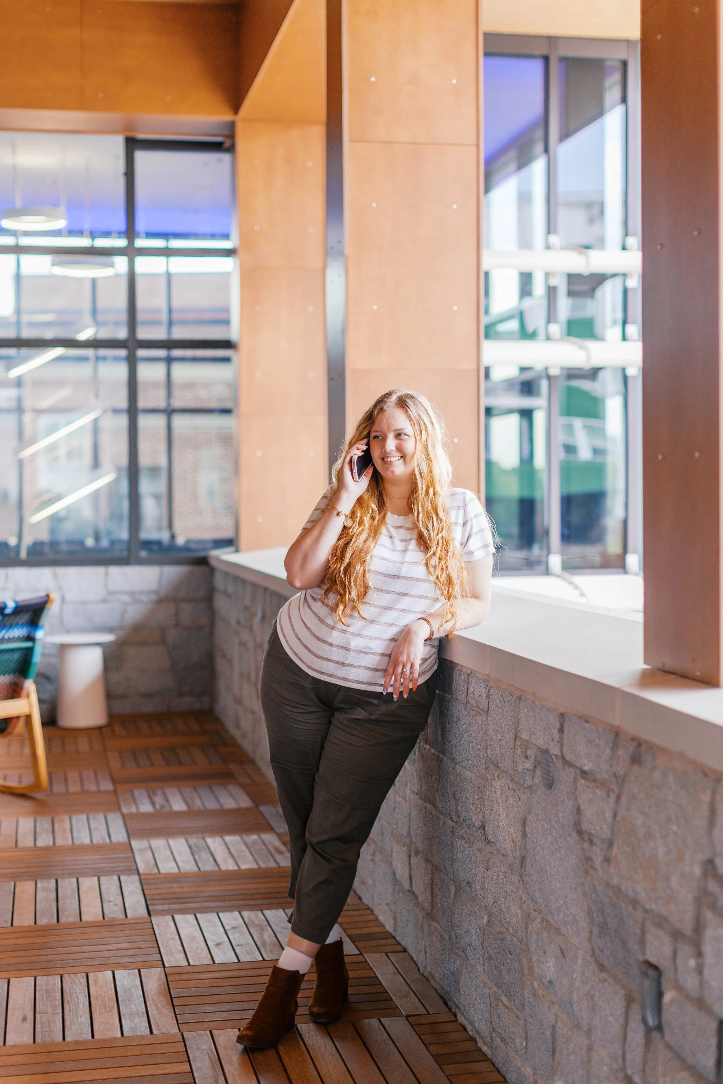 A woman with long red hair, wearing a striped t-shirt, dark pants, and brown boots, is smiling while talking on her cell phone near large windows with a city view.