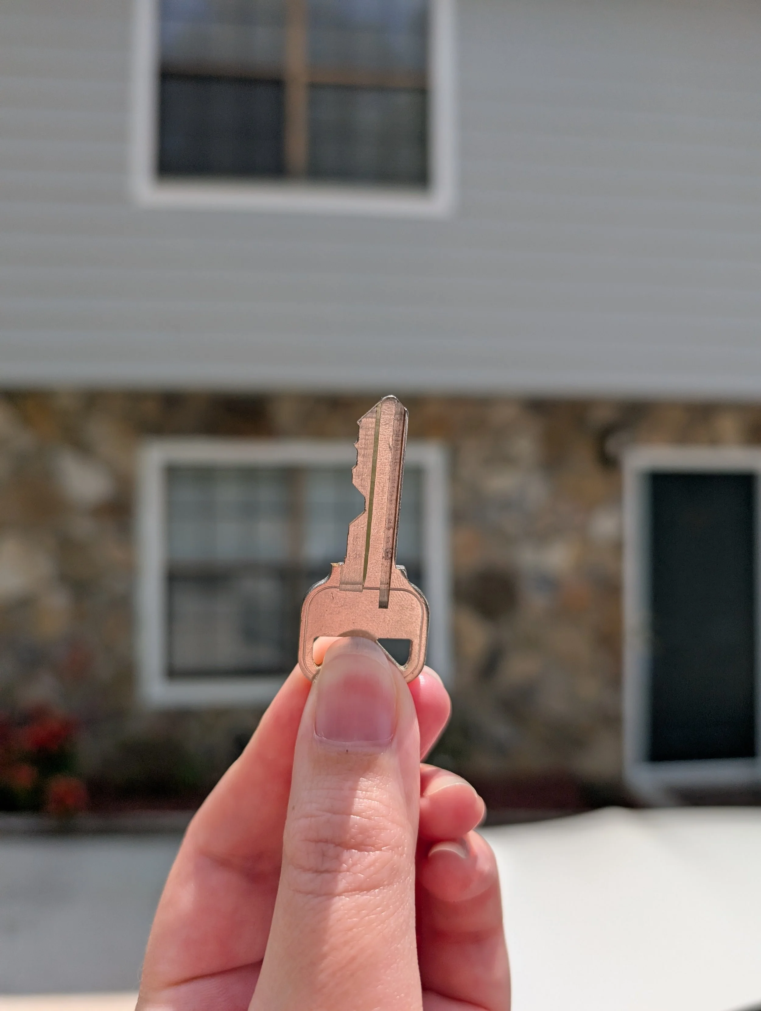 Close-up of a person's hand holding a silver key in front of a house with a stone and siding exterior and multiple windows.