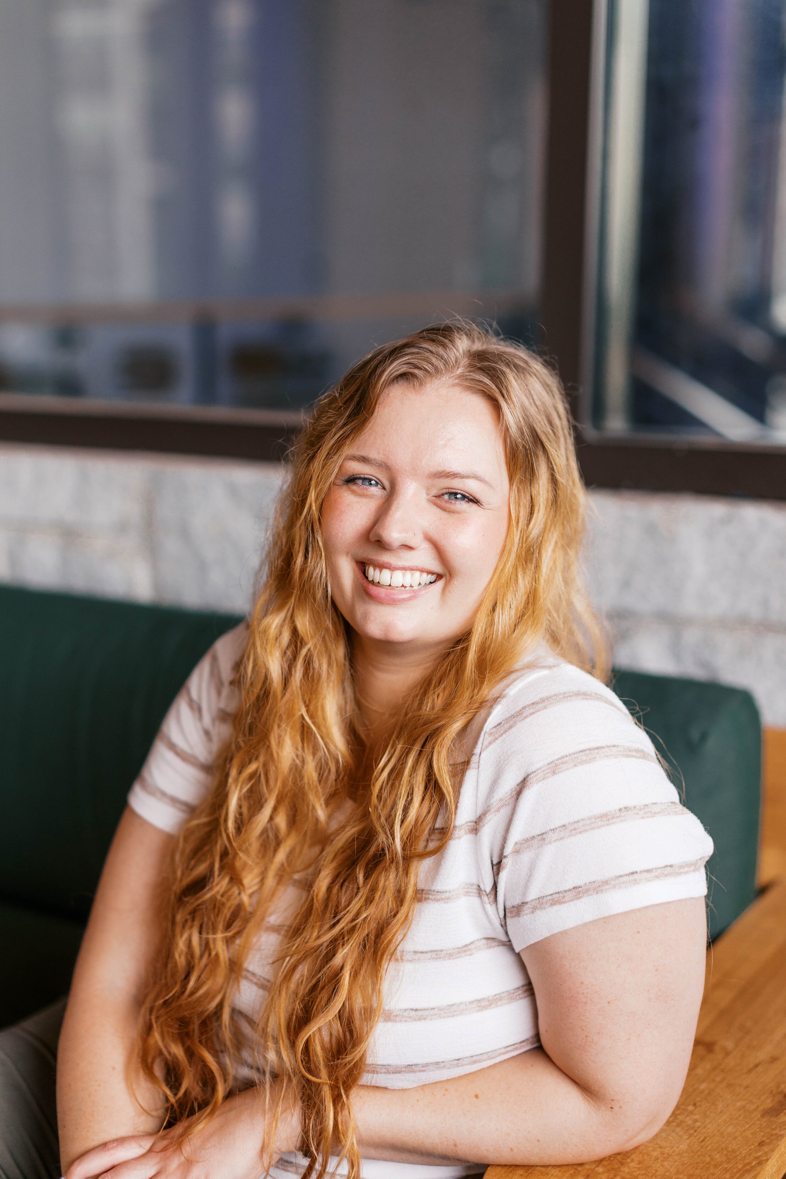 A smiling woman with long red hair, wearing a striped t-shirt, sitting in a casual setting with a window behind her.