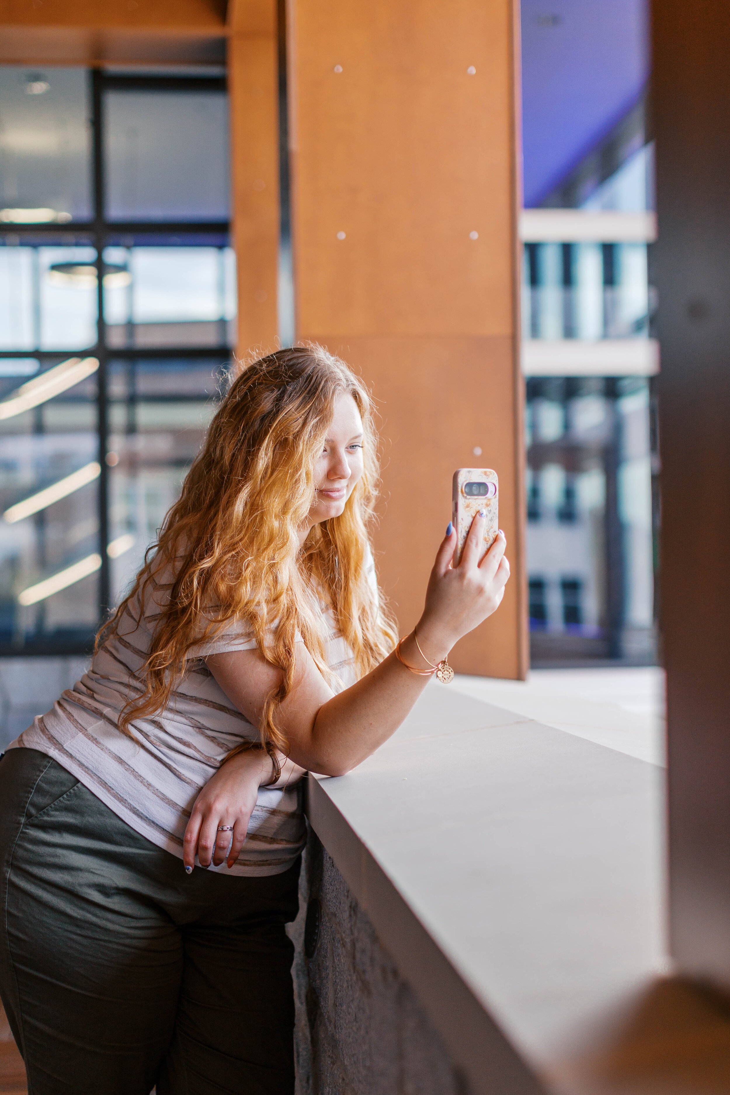 A young woman with long, curly red hair taking a selfie with her smartphone inside a modern building with large windows.