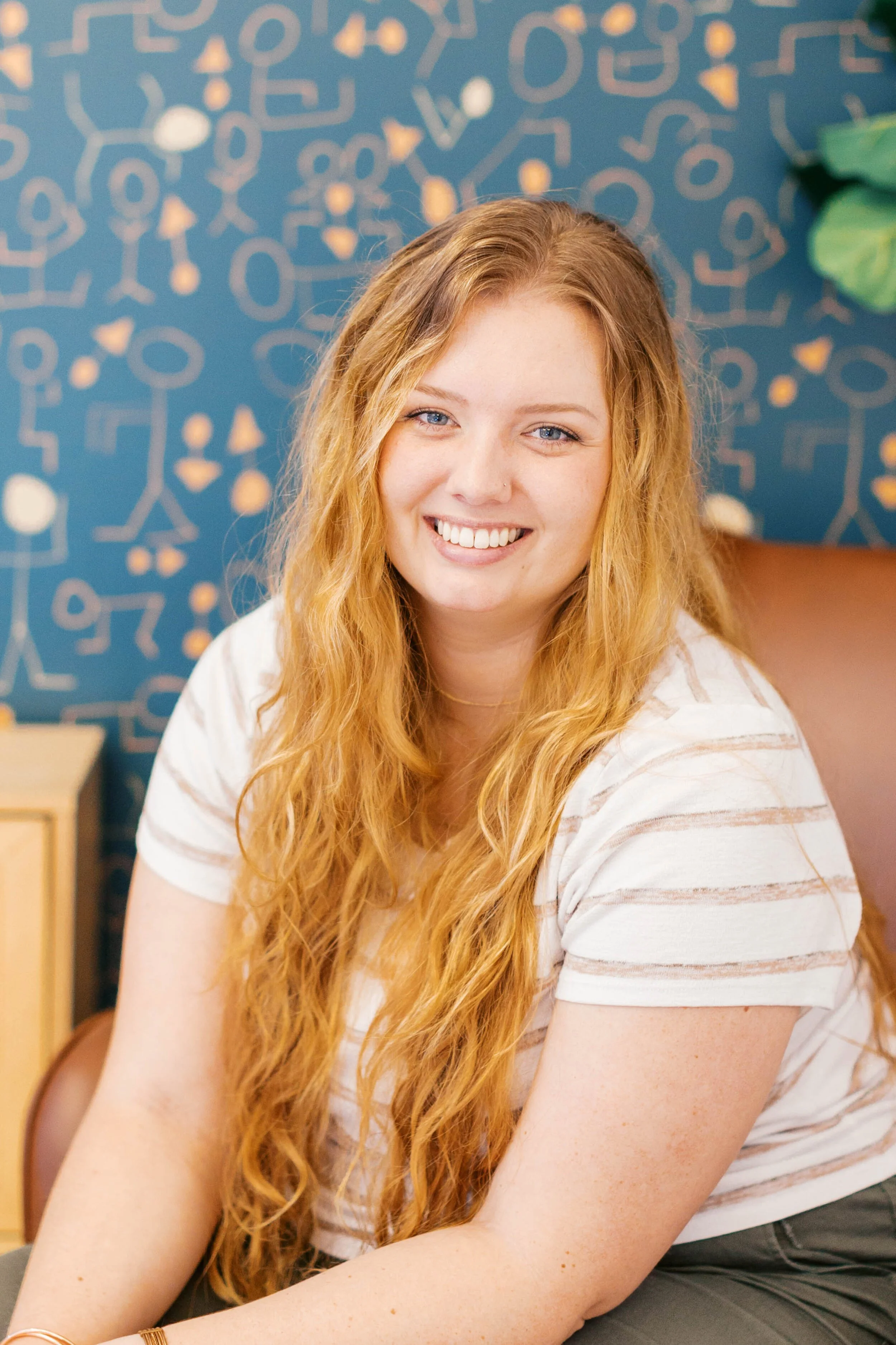 A young woman with long curly red hair, smiling, sitting indoors with a patterned blue wall in the background.