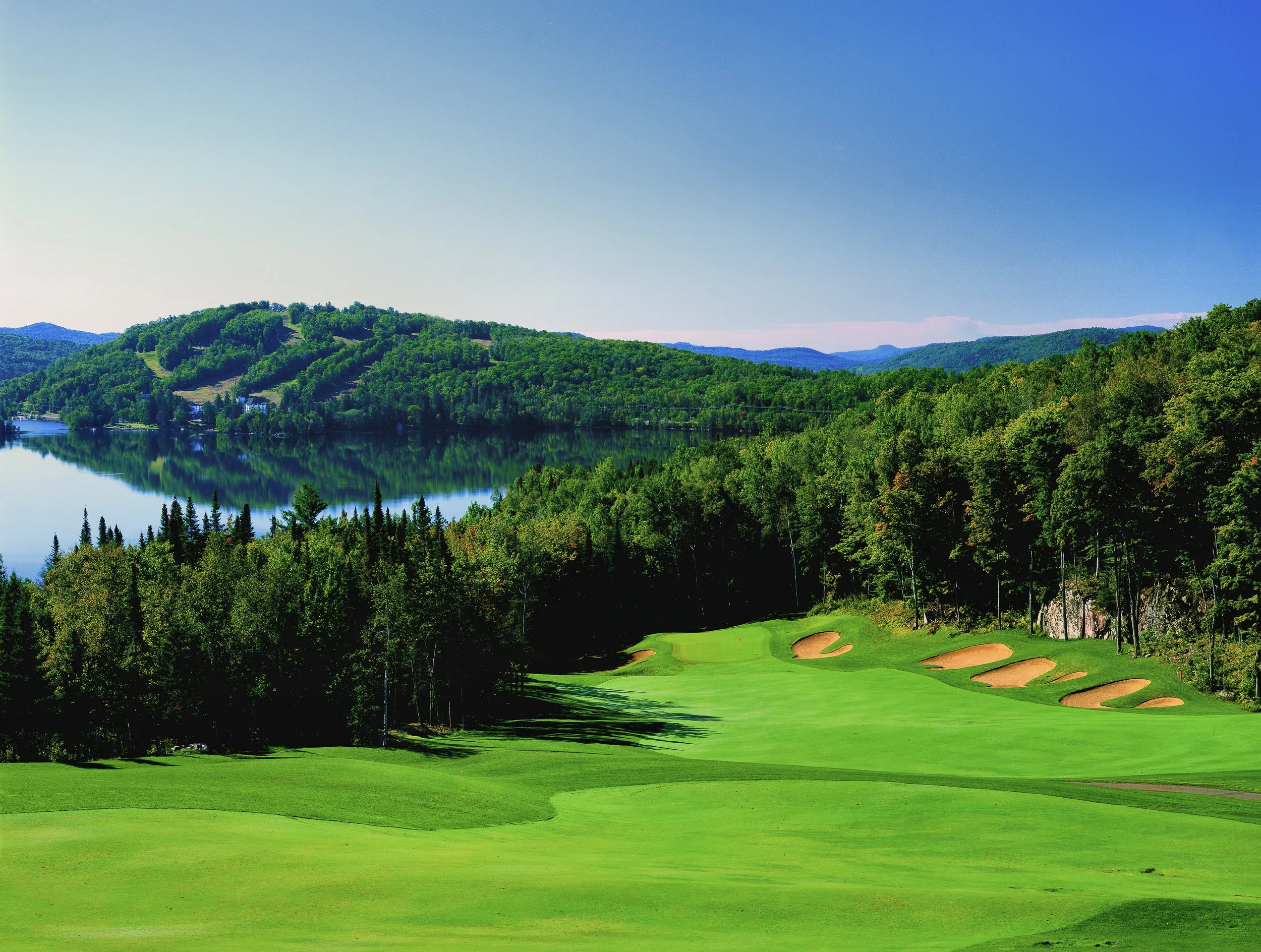 Scenic view of a golf course with sand bunkers, surrounded by lush forests and a reflective lake.