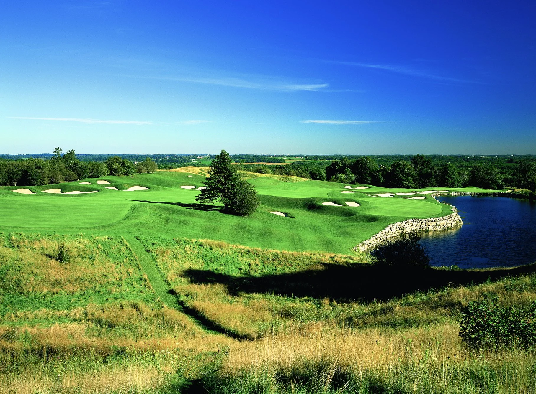 Golf course landscape with green fairways, sand bunkers, trees, and a pond under a clear blue sky.