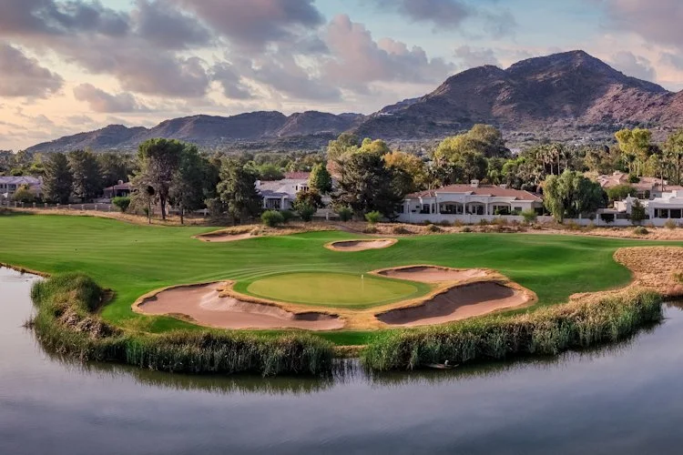 Scenic golf course with a green surrounded by sand bunkers, bordered by water, with residential houses and mountains in the background.
