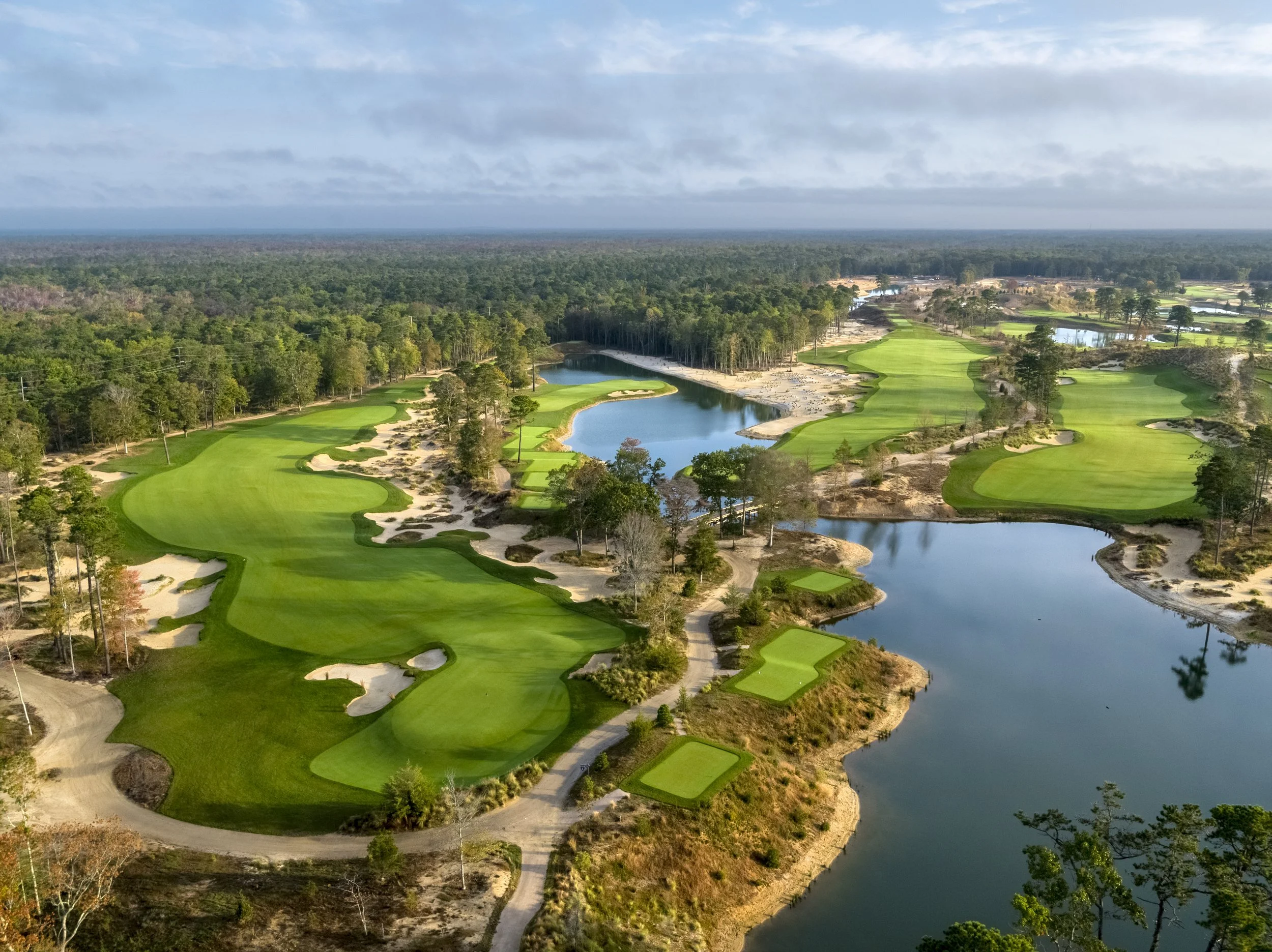 Aerial view of a scenic golf course with green fairways, sand traps, water hazards, and surrounding trees under a blue sky.