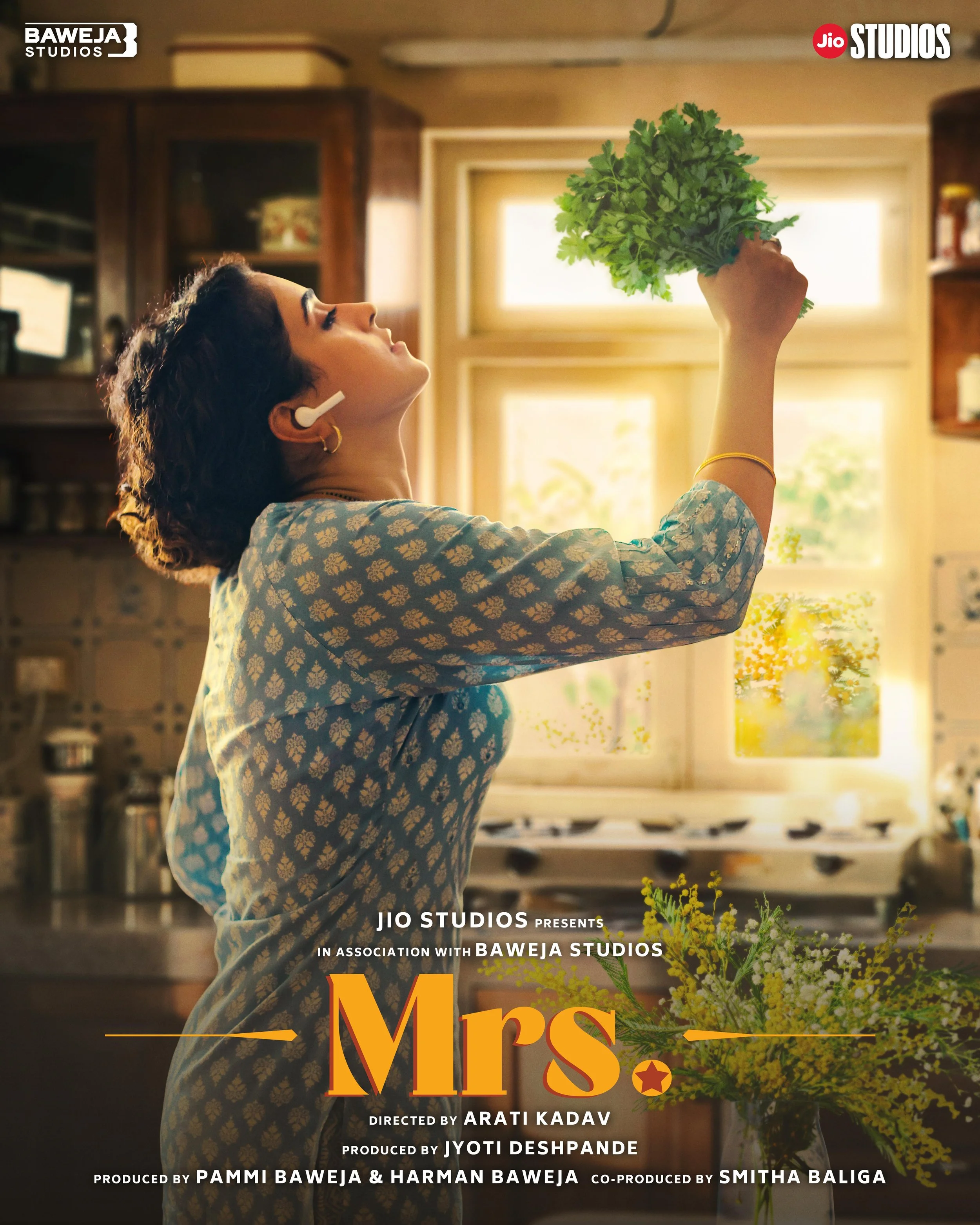 A woman in a patterned blue dress is holding a bunch of green leafy vegetables in her right hand, in a kitchen with sunlight coming through the windows. She is wearing wireless earbuds and has short, curly hair.