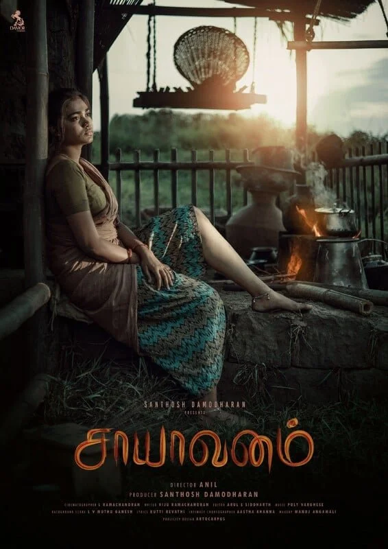 A woman sitting on a traditional woven mat next to an outdoor cooking stove with a small fire, set against a rural landscape at sunset with a thatched roof and hanging basket.