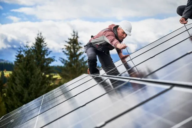 A person wearing a white helmet and a pink jacket working on solar panels outdoors with trees and blue sky in the background.
