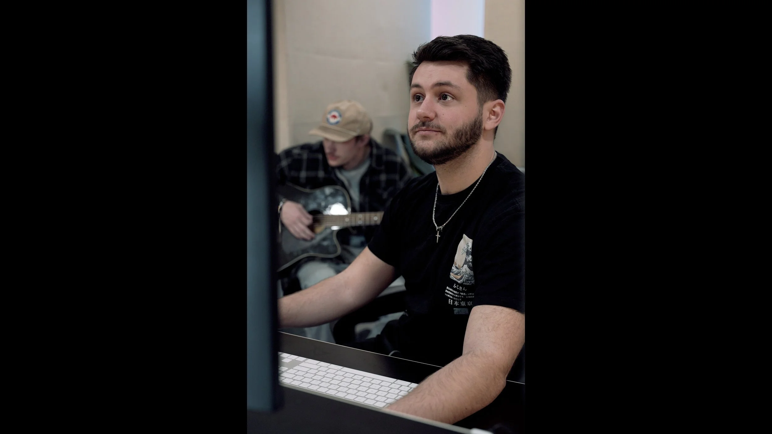 A young man with dark hair and a beard sitting at a computer with a keyboard, looking to the side. In the background, another person wearing a beige cap and plaid shirt is playing an acoustic guitar.