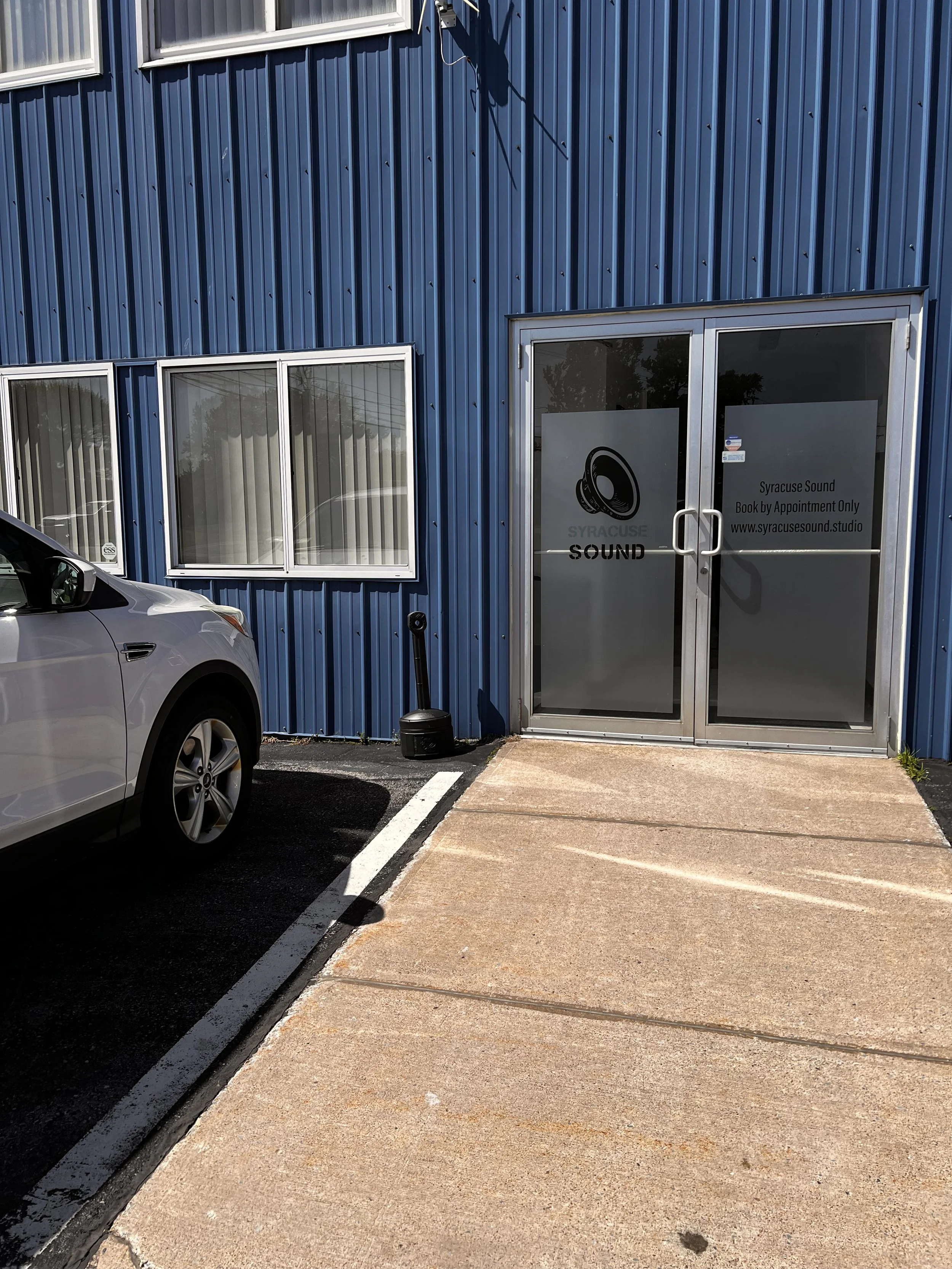 Blue industrial building with vertical metal siding, three windows, a glass door with signage for Syracuse Sound, a parked white vehicle, and a concrete sidewalk and parking lot.