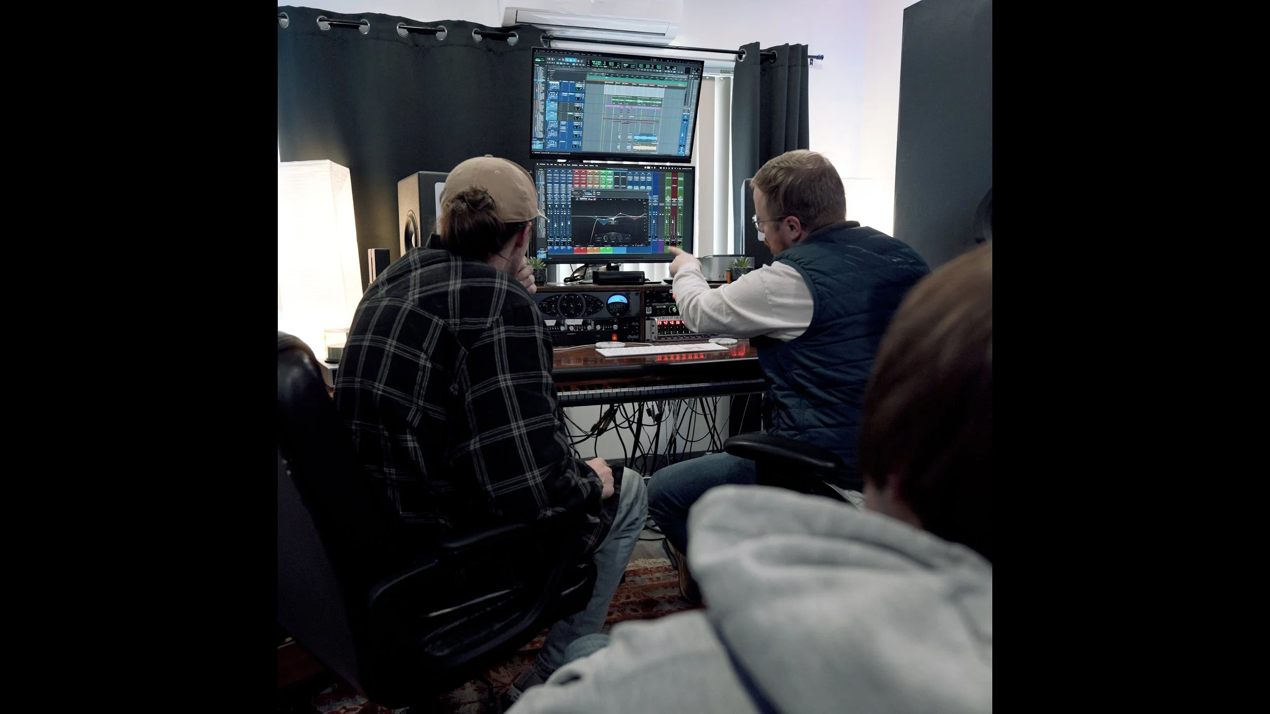 Two men sitting in front of computer monitors displaying digital audio workstation software, working on music production in a studio.
