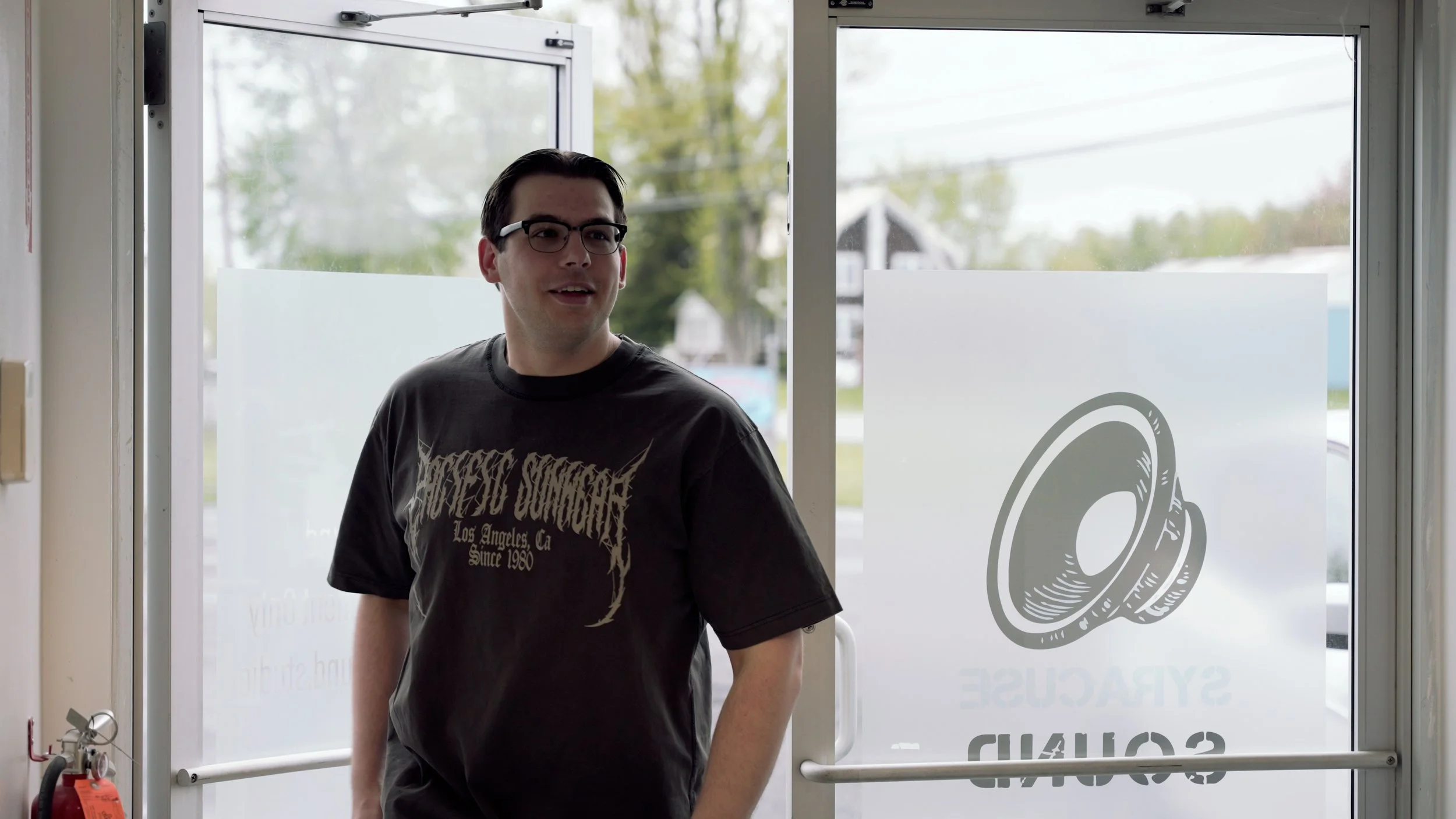 A young man with dark hair and glasses standing inside near glass doors with a tired-looking expression, wearing a black T-shirt with white text. Outside the doors, trees and houses are visible.