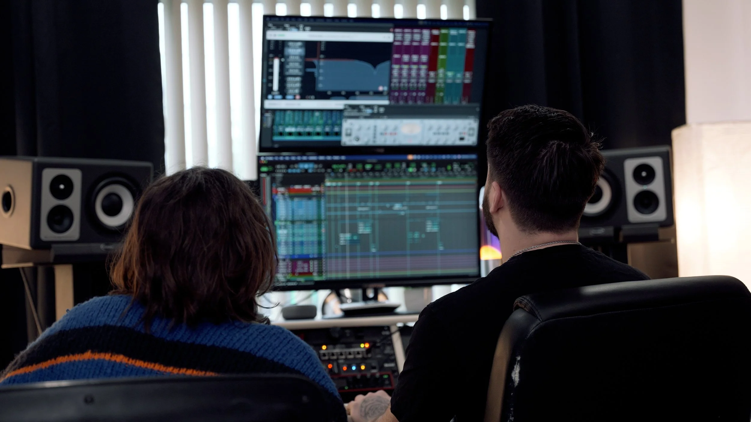 Two people sitting in front of computer screens displaying music production software and audio editing tracks, with speakers on either side of the screens.