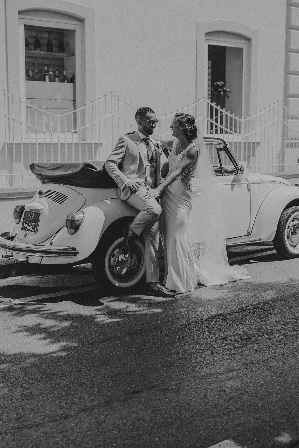 A bride and groom in wedding attire smiling and holding hands, leaning against a vintage car on a street during the day.