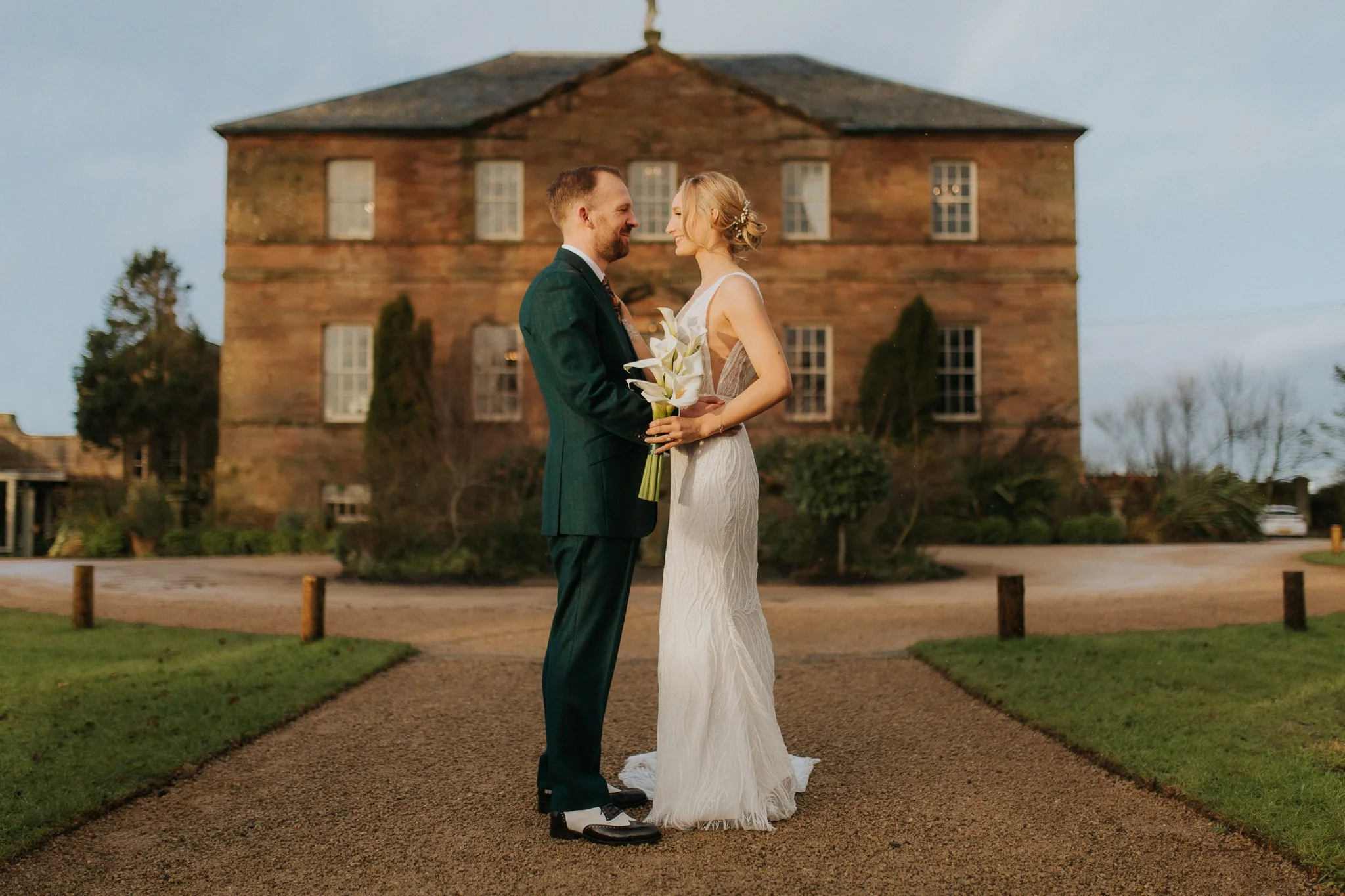 A bride and groom standing close together outdoors during sunset, smiling at each other, holding a bouquet of white lilies, with a historic brick building in the background.