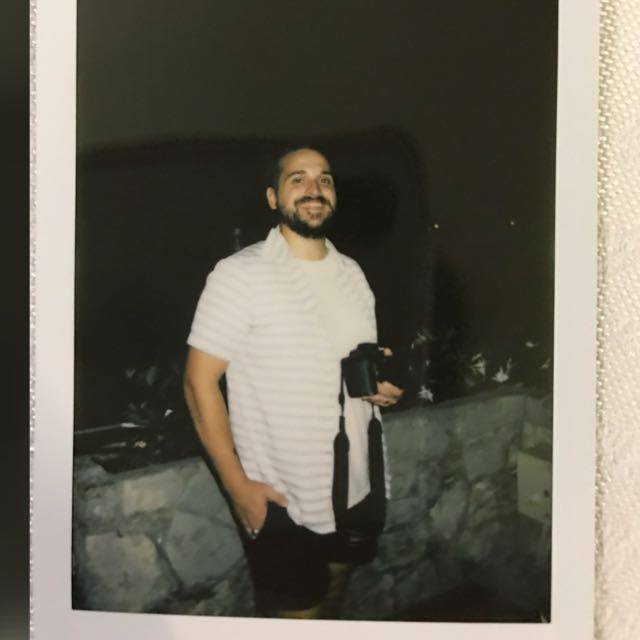 A man with a beard and smiling face, wearing a light-colored striped shirt, is standing outdoors at night, holding a camera in his right hand, and posing next to a low stone wall.