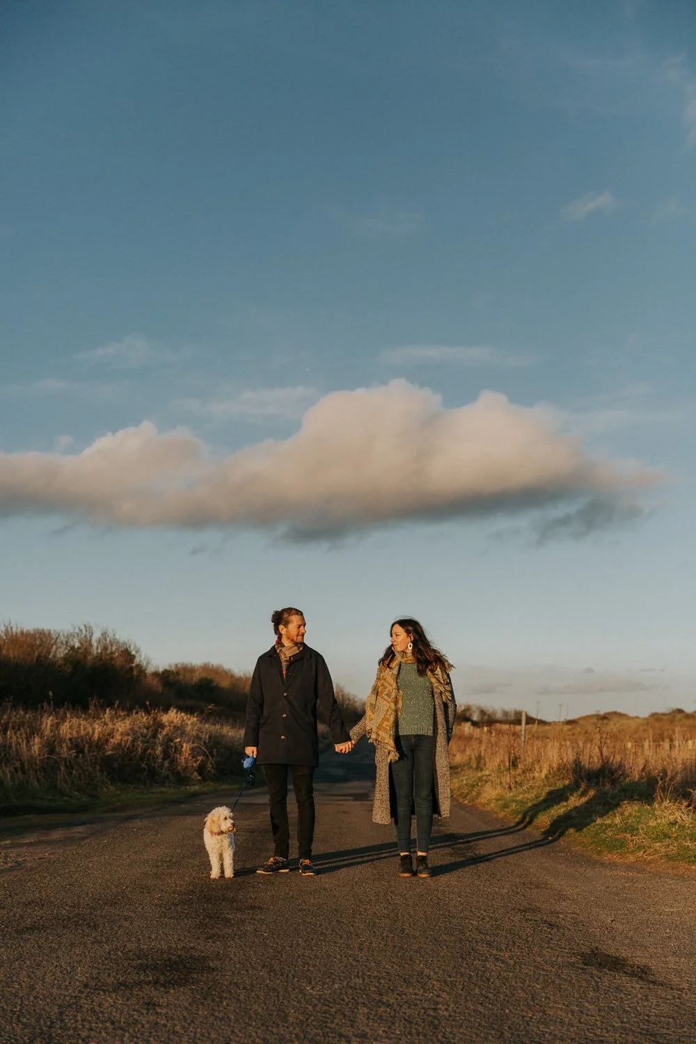 A couple holding hands with a dog walking on a rural road during sunset.